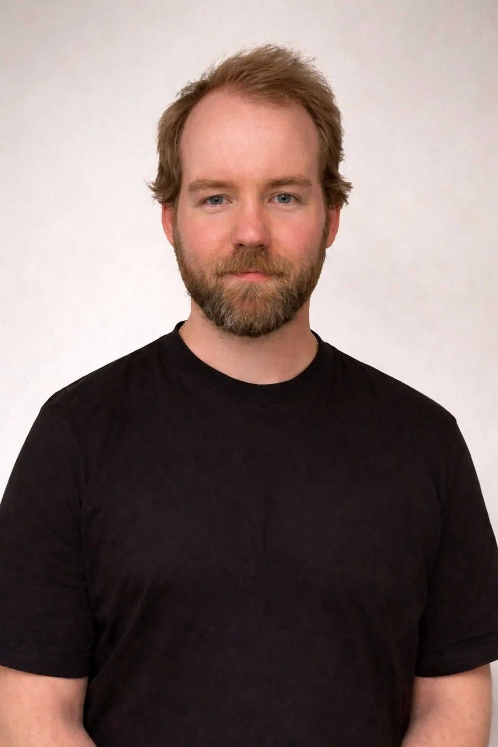 A man with light skin, blue eyes, a beard, and medium-length brown hair is standing against a plain white background, wearing a black t-shirt.