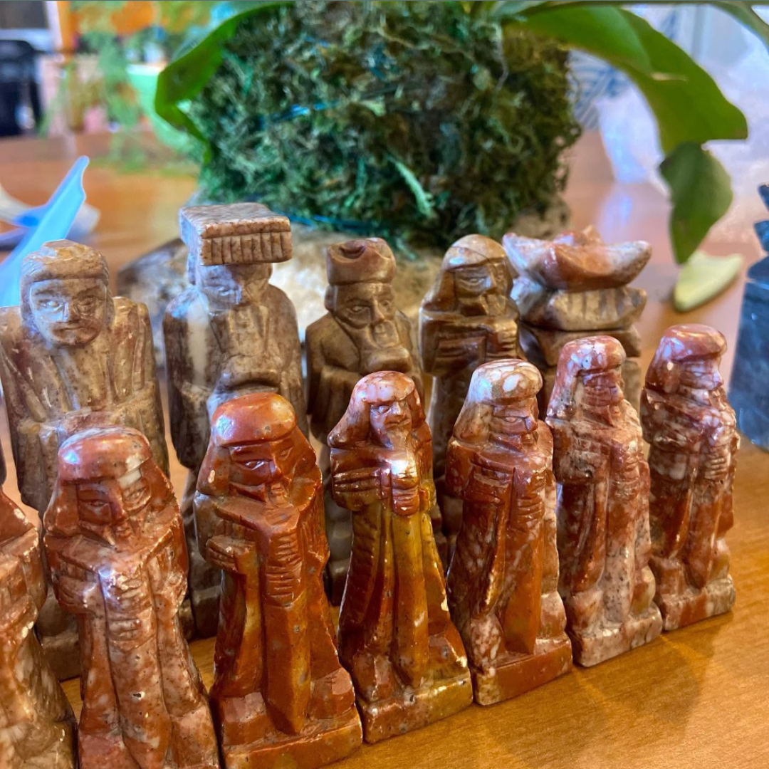 Collection of carved stone figurines with detailed facial features and traditional clothing, placed on a wooden surface with a blurred background of plants and other objects.