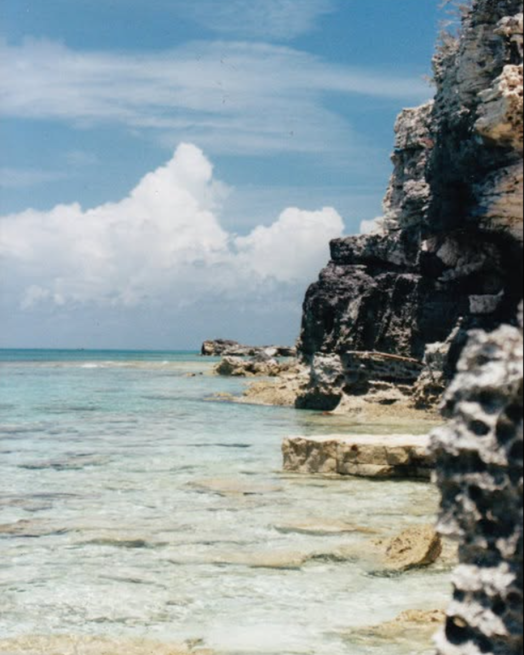 A rocky beach with clear turquoise water and a partly cloudy sky.