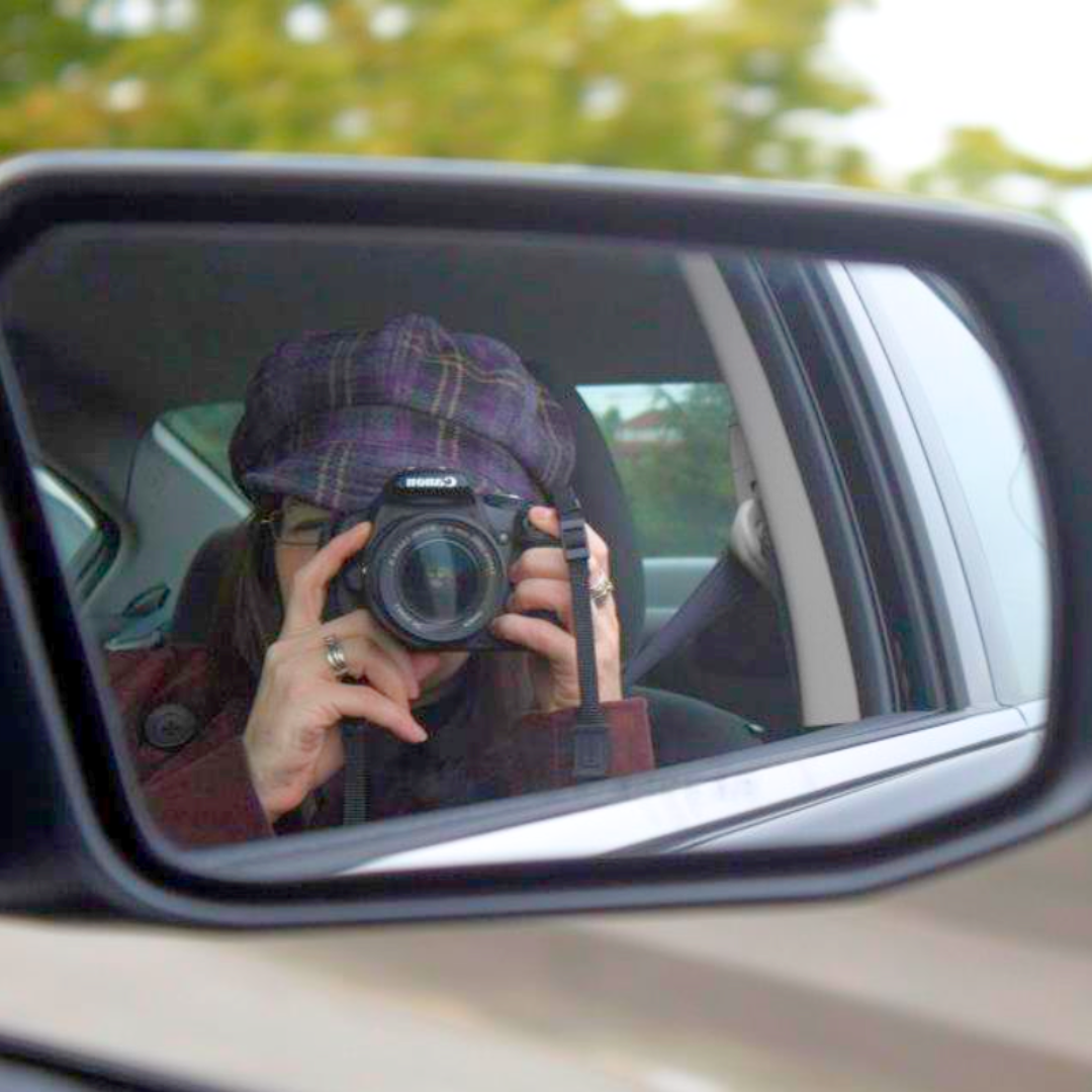 Person taking a photo with a camera in a car's side mirror, wearing a plaid hat and rings.
