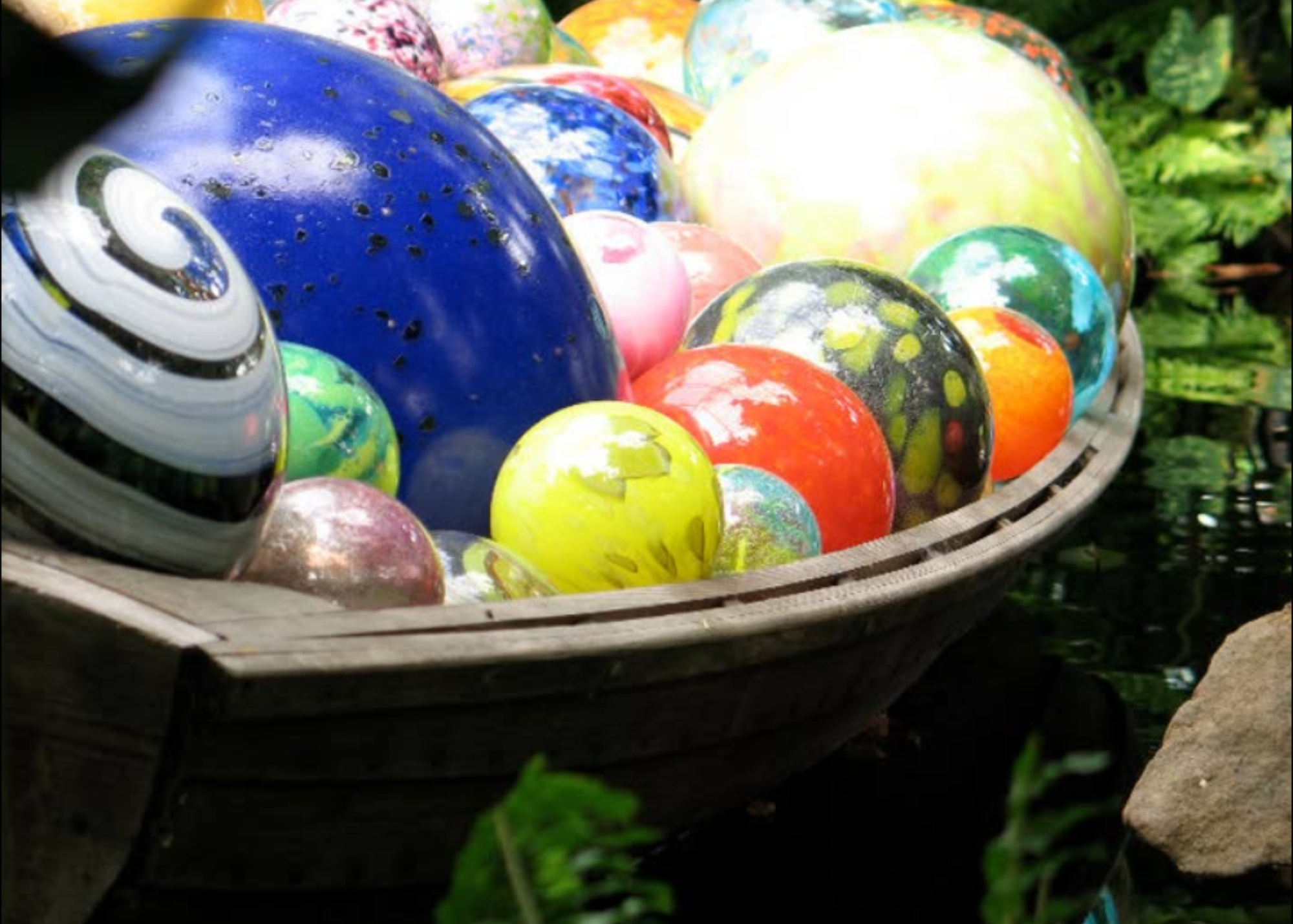 Colorful glass marbles in a basket outdoors with green grass and plants surrounding it.