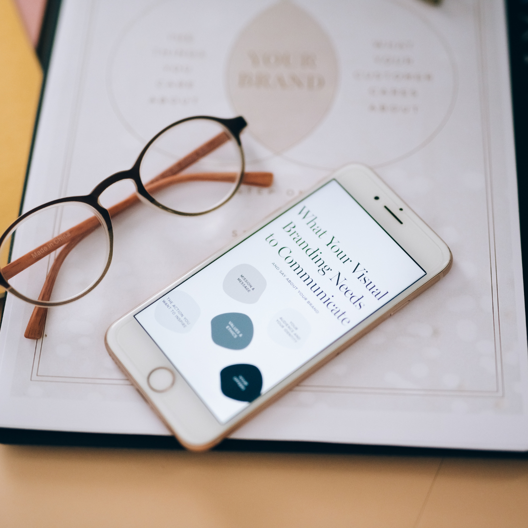 A smartphone displaying a webpage with the title 'What Your Visual Branding Needs to Communicate' placed on a magazine or brochure on a desk. A pair of round eyeglasses and a brown pencil are also resting on the magazine.