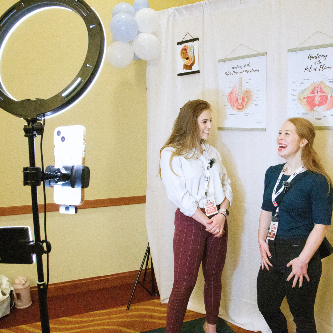 Two women are standing and talking in front of a curtain with anatomical posters on the wall, while being recorded by a ring light and a smartphone mounted on a tripod. One woman is wearing a white blouse and red checkered pants, the other is in a dark blue top and black pants.