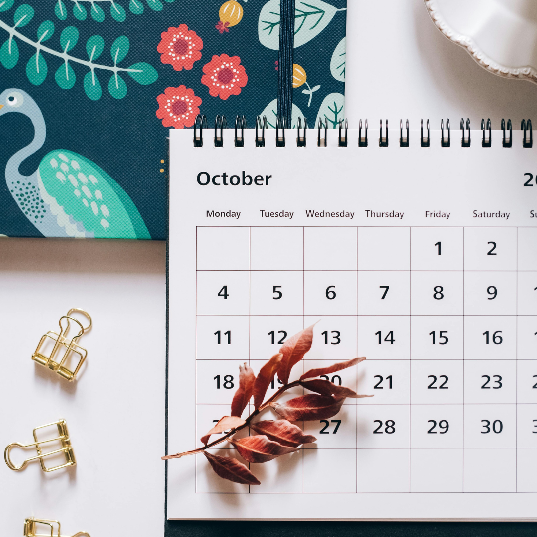 A desk calendar open to October with a dried branch of leaves resting on the date 13. To the left are three gold binder clips, and in the top right corner is part of a white plate. In the background is a patterned fabric with a teal and white peacock and floral design.
