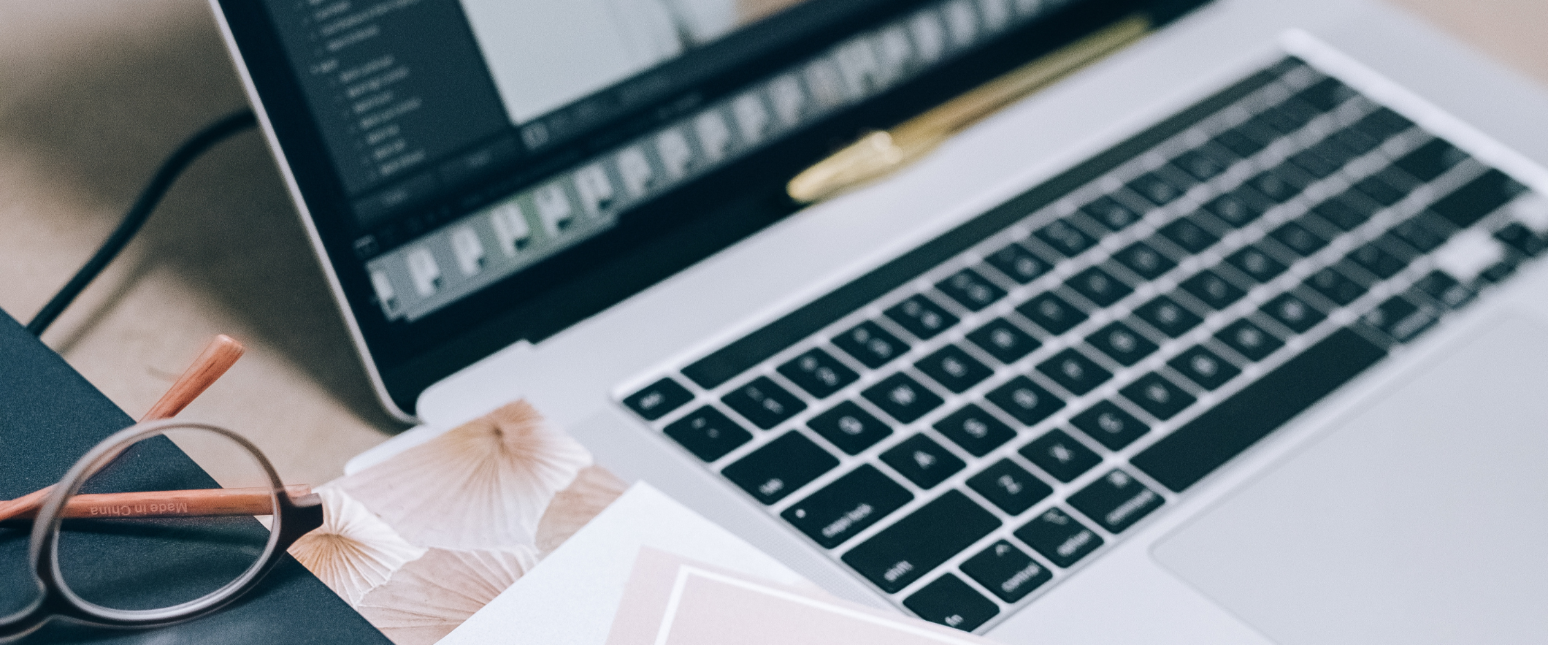 Close-up of a laptop keyboard and screen, with reading glasses, a notebook, and a pen on a wooden desk.