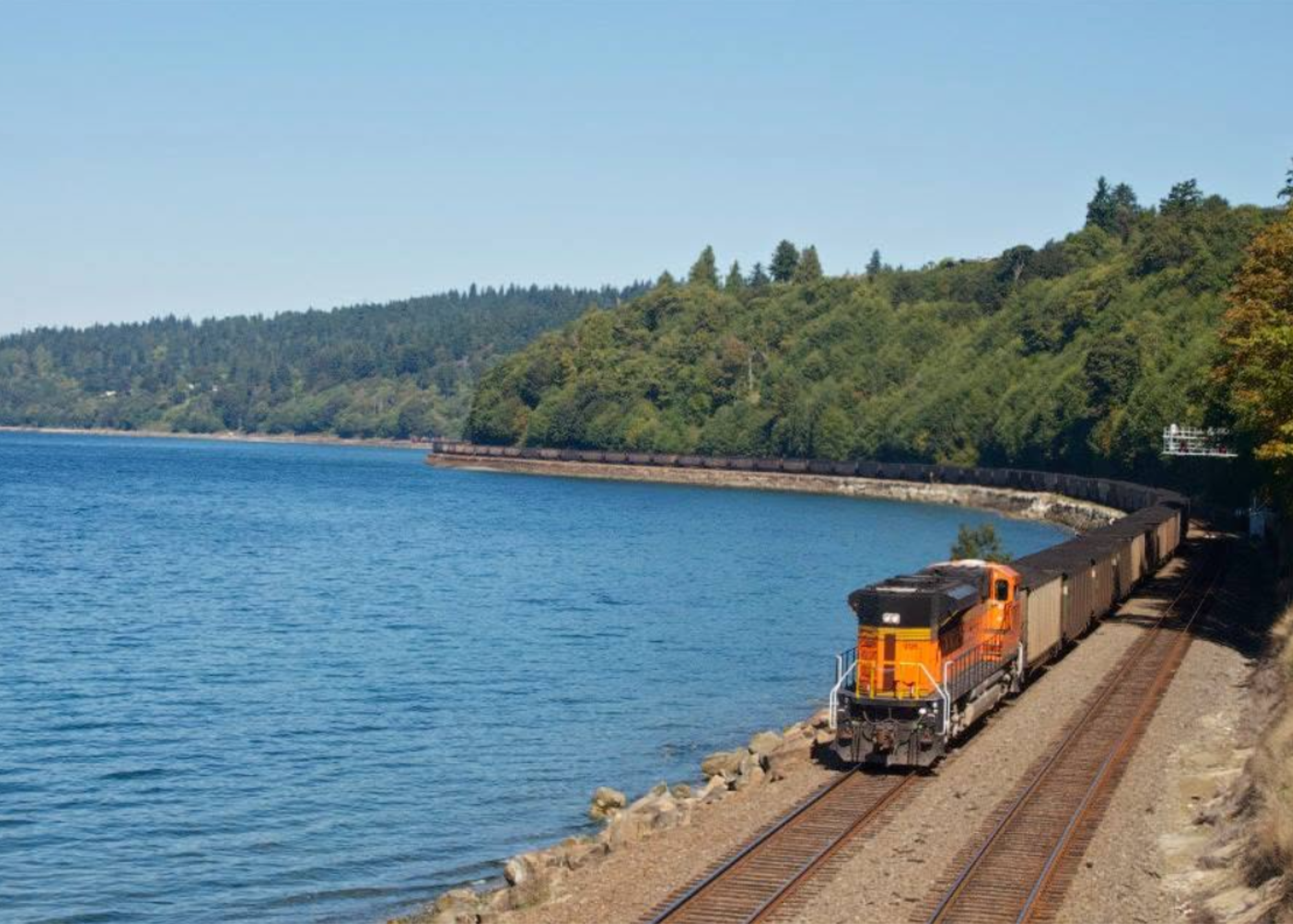A train running along a track beside a large body of water with a forested hillside in the background.