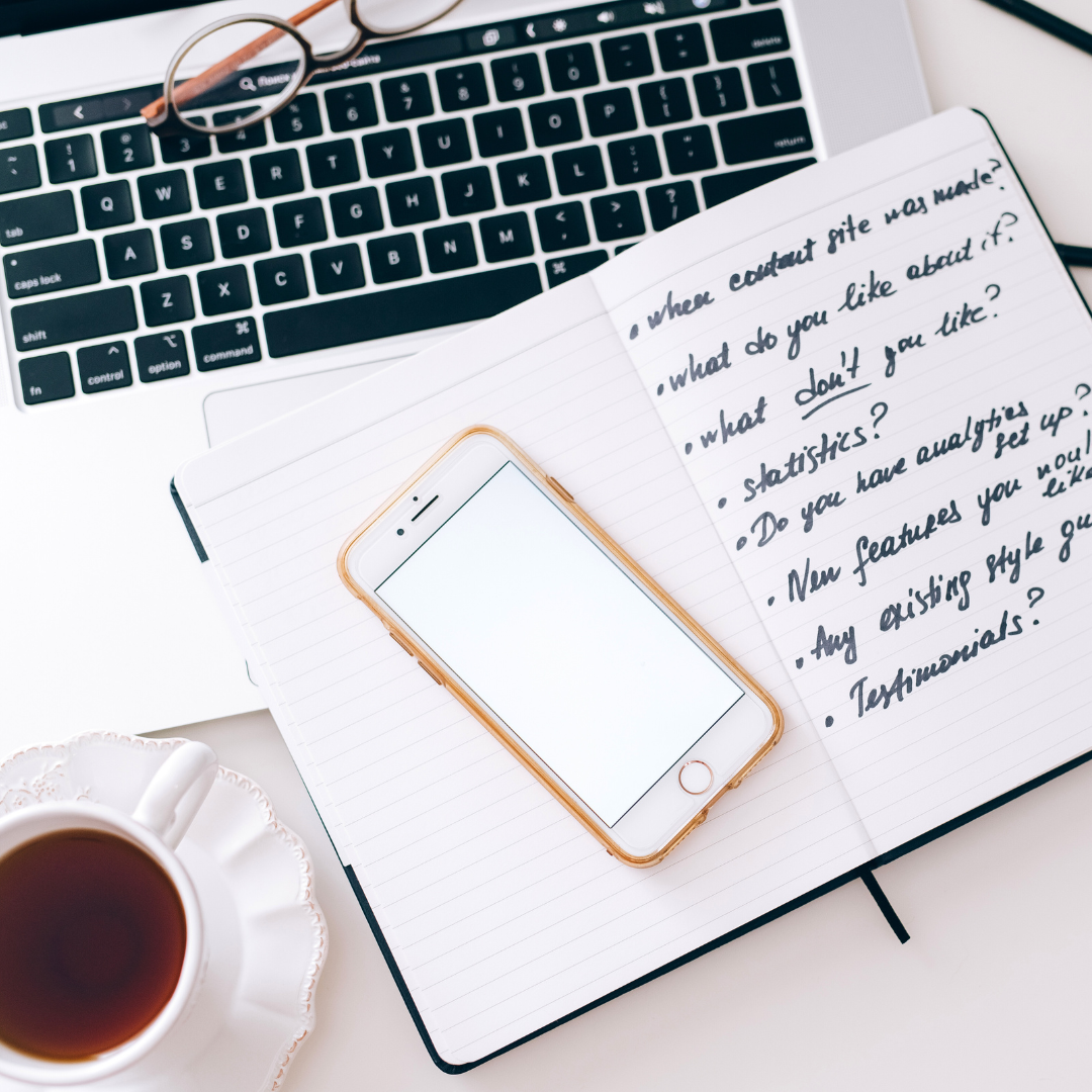 A workspace with a laptop keyboard, a notebook with handwritten notes, a smartphone, a cup of tea, and a pencil.