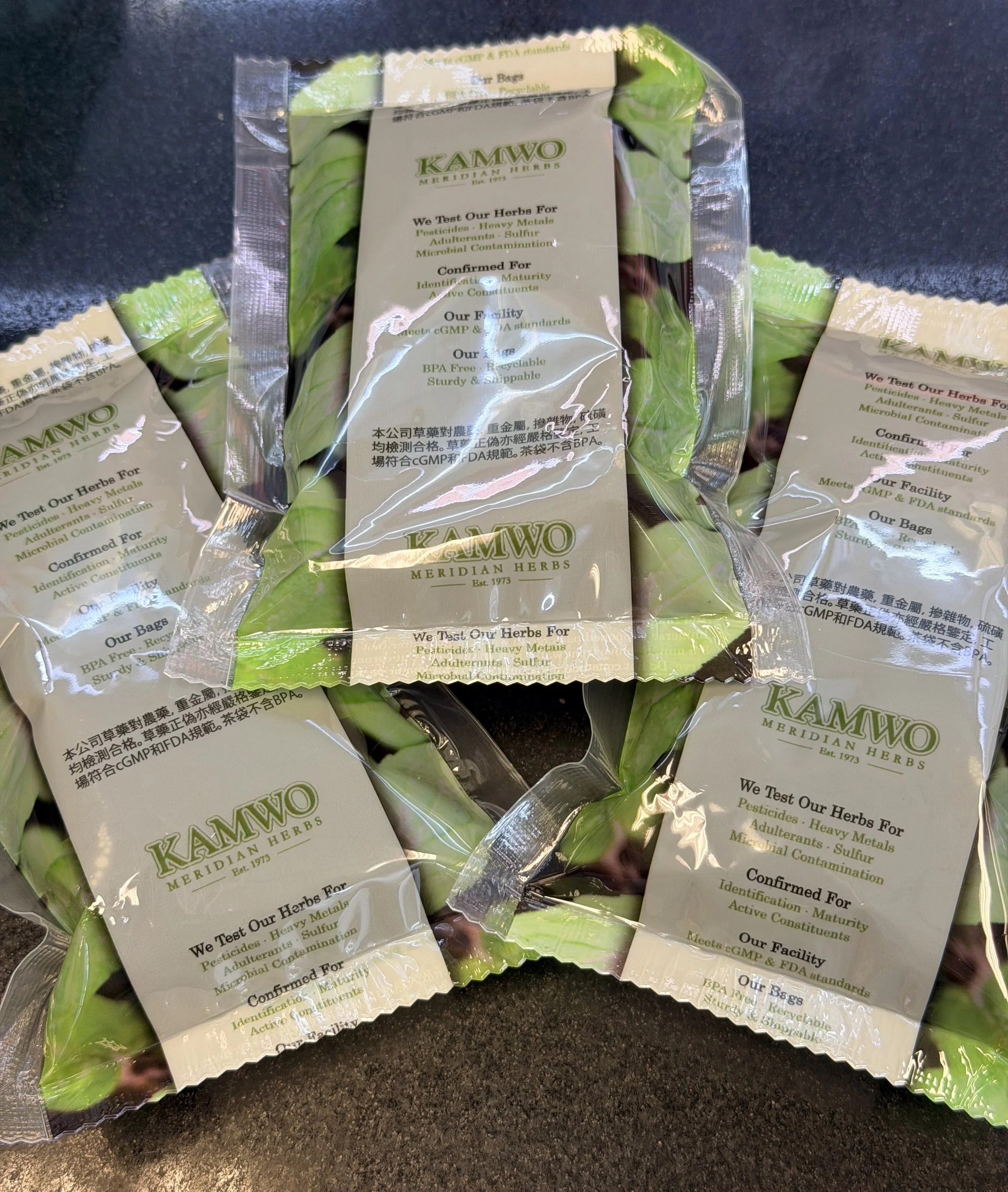 Multiple small packages of Kamwo Meridian Herbs with labels indicating testing for pesticides, heavy metals, adulterants, sulfur, and microbial contamination, and confirming for identification, maturity, and active constituents.