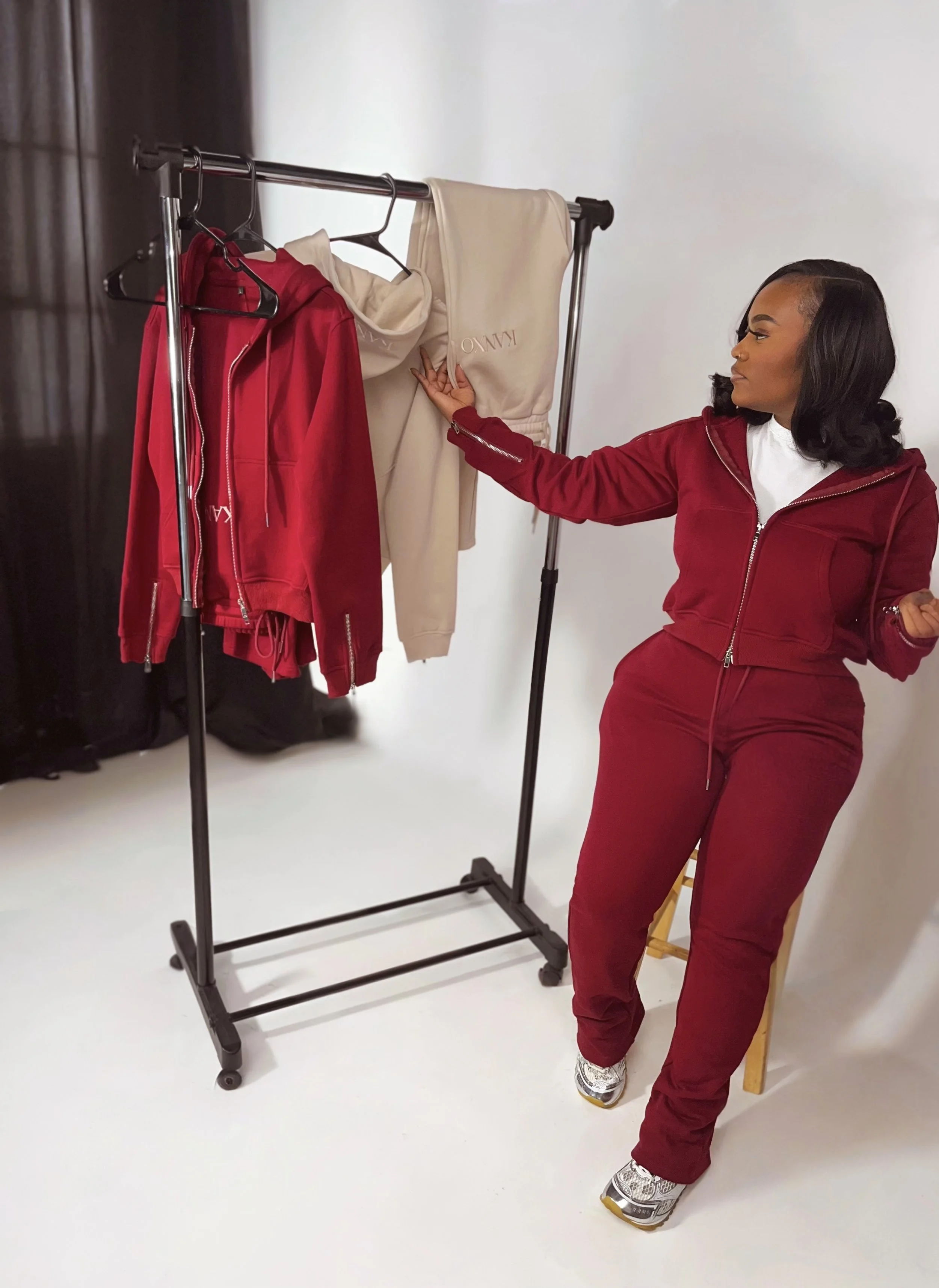 A woman in a matching red hoodie and sweatpants stands next to a clothing rack with red and beige hoodies and sweatpants hanging on it.