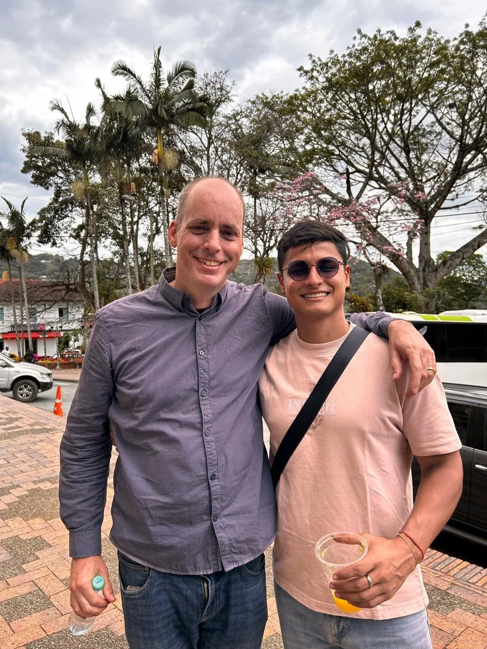 Two men smiling and standing close together outdoors, one wearing glasses and a pink T-shirt, holding a drink, the other in a gray shirt, with trees and parked cars in the background.