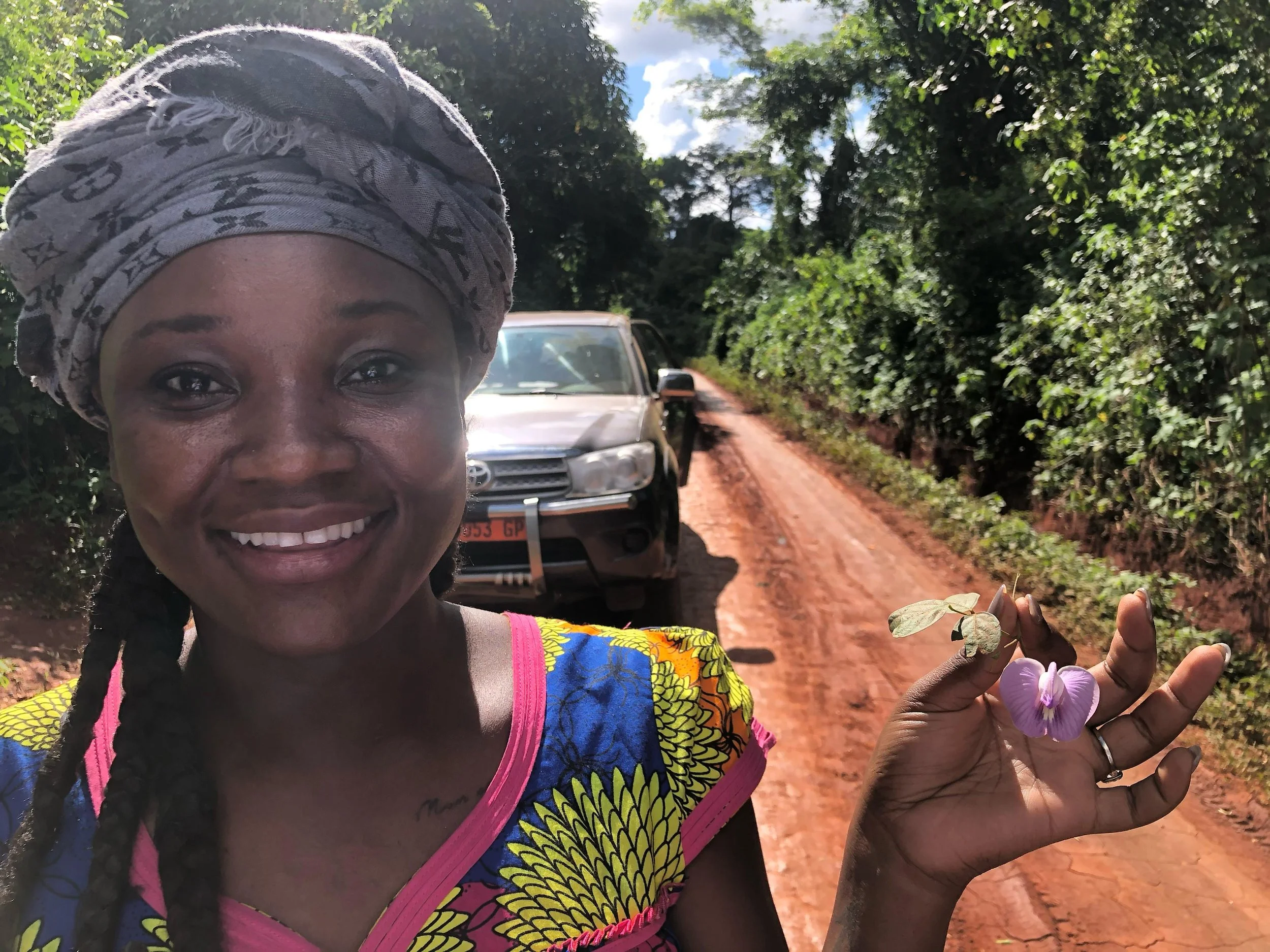 Woman smiling on a dirt road holding a butterfly-shaped flower, with a car and green foliage in the background.