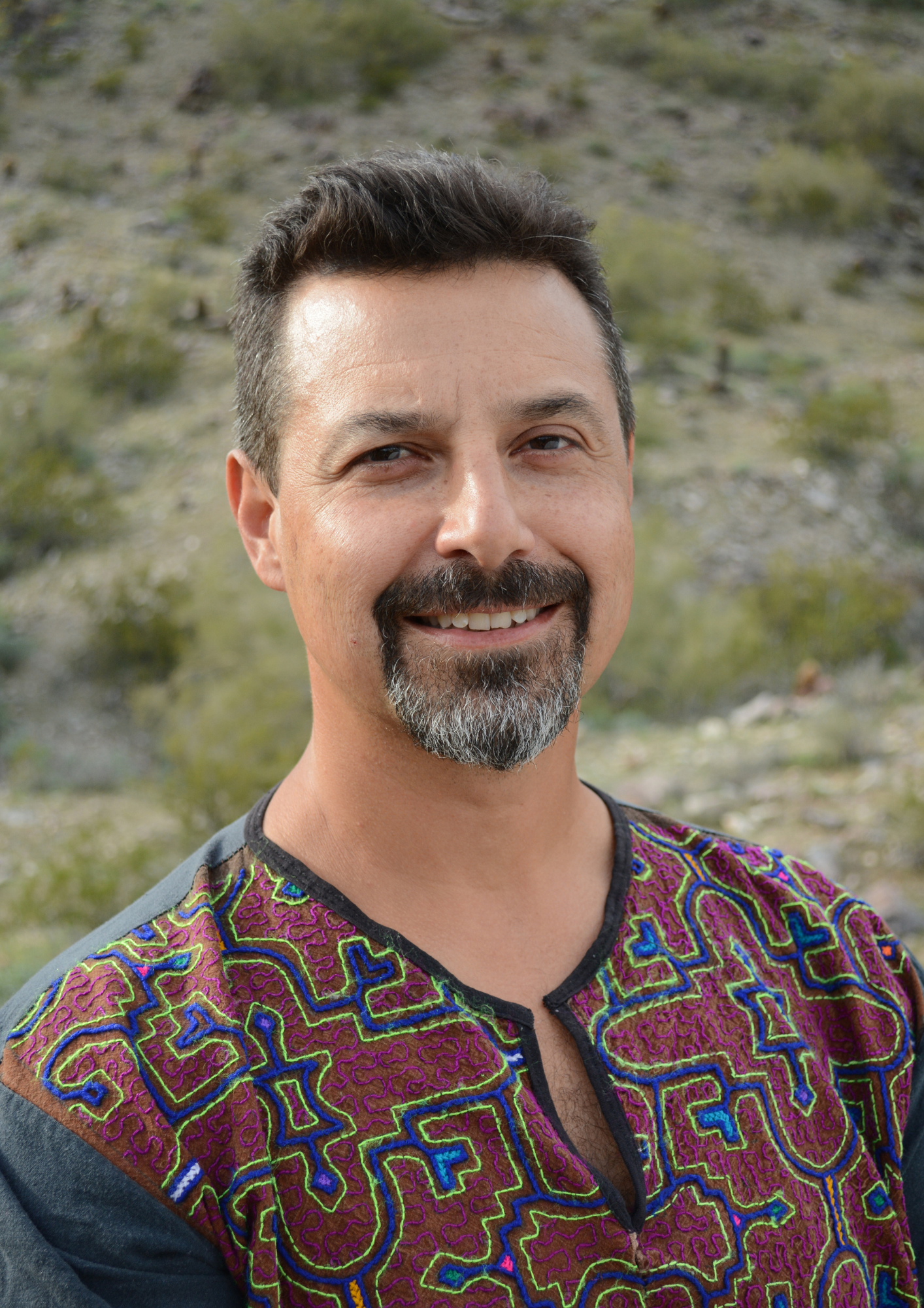 A man with dark hair, a beard, and gray highlights standing outdoors with a mountainous background, wearing a colorful embroidered shirt.