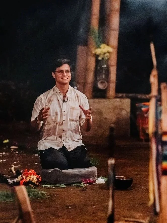 A person kneeling on the ground, smiling and gesturing with hands, amidst various offerings and flowers, in an outdoor, possibly spiritual or ritual setting.