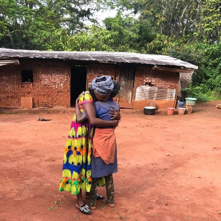 Two women, one in a colorful patterned dress and the other in a blue dress with an orange cloth, hugging outside a rustic brick house with a tin roof and various containers nearby.
