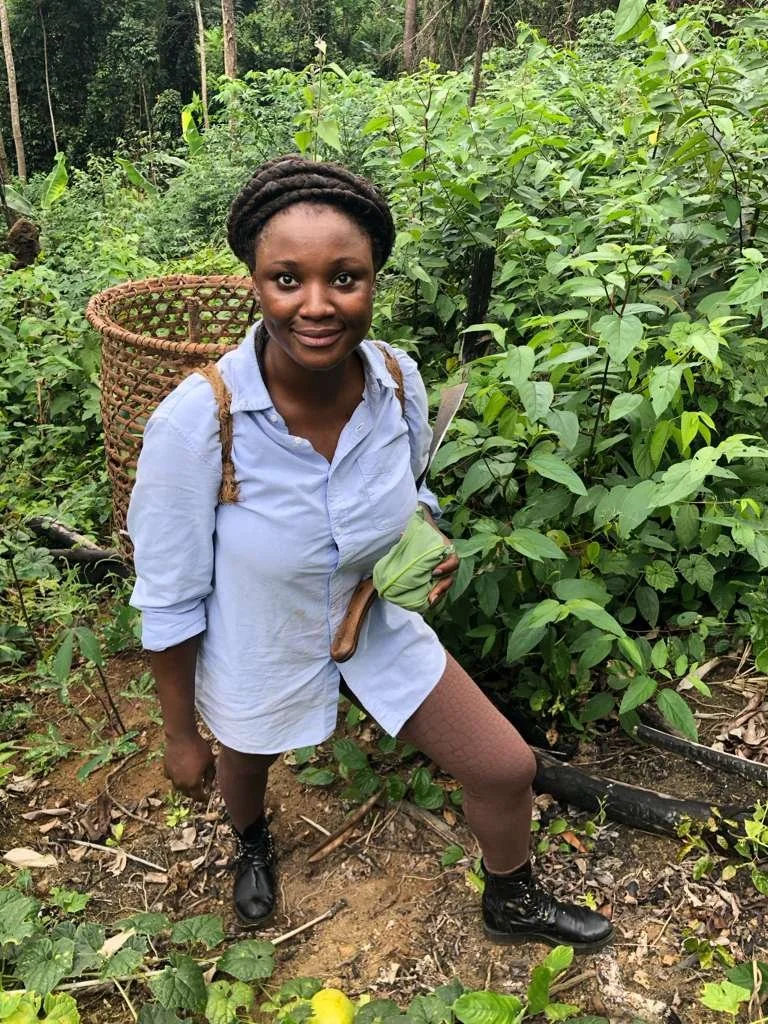 A young woman with dark curly hair in an outdoor green garden or forest, holding a knife and some green leaves, wearing a blue shirt, fishnet stockings, black shoes, and carrying a large wicker basket on her back.