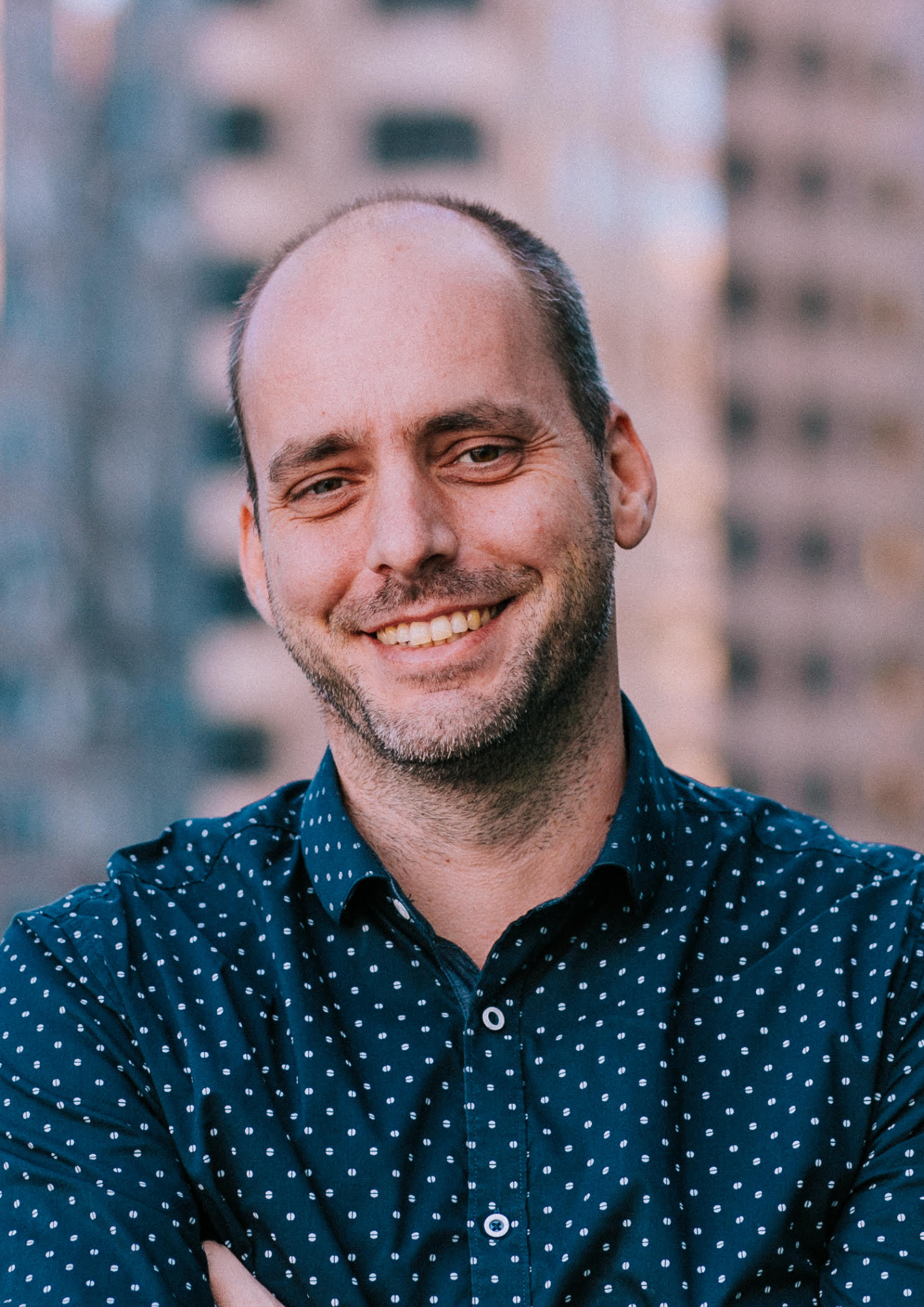 Smiling man with a beard and receding hairline, wearing a navy blue, polka-dotted shirt, standing outdoors with city buildings in the background.
