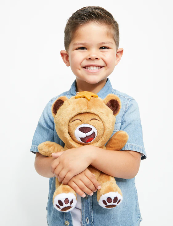 A young boy with short hair smiling and hugging a plush toy of a cartoon bear with a yellow crown on its head.