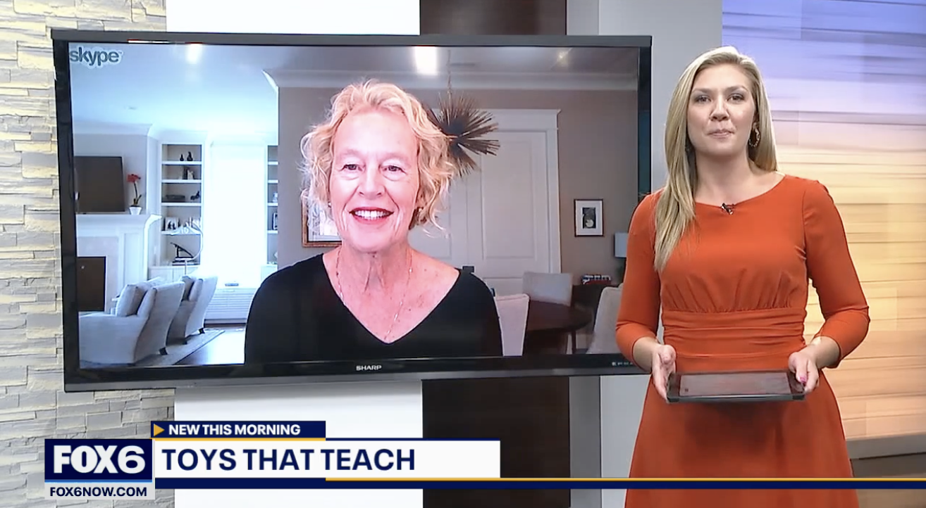 A female news anchor standing in a studio holding a tablet, with a large screen behind her displaying a video call with an older woman in a living room setting.