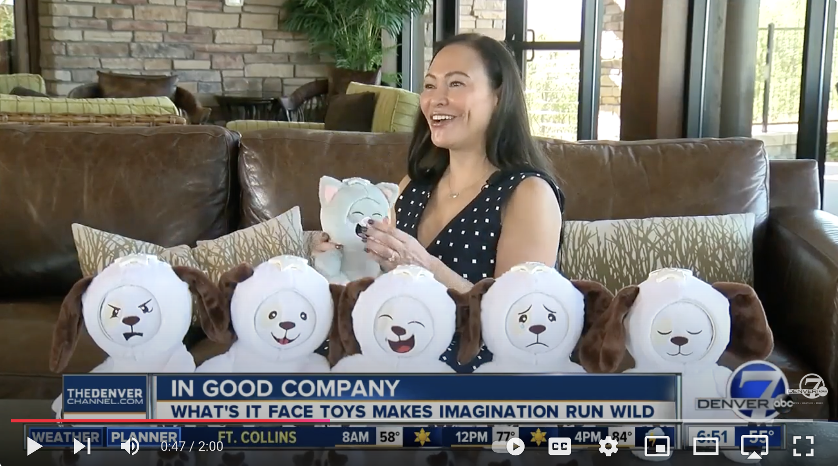 Woman sitting on a brown leather couch holding a plush toy dog, surrounded by six plush toy dogs with various facial expressions, in a setting with large windows and a brick wall.