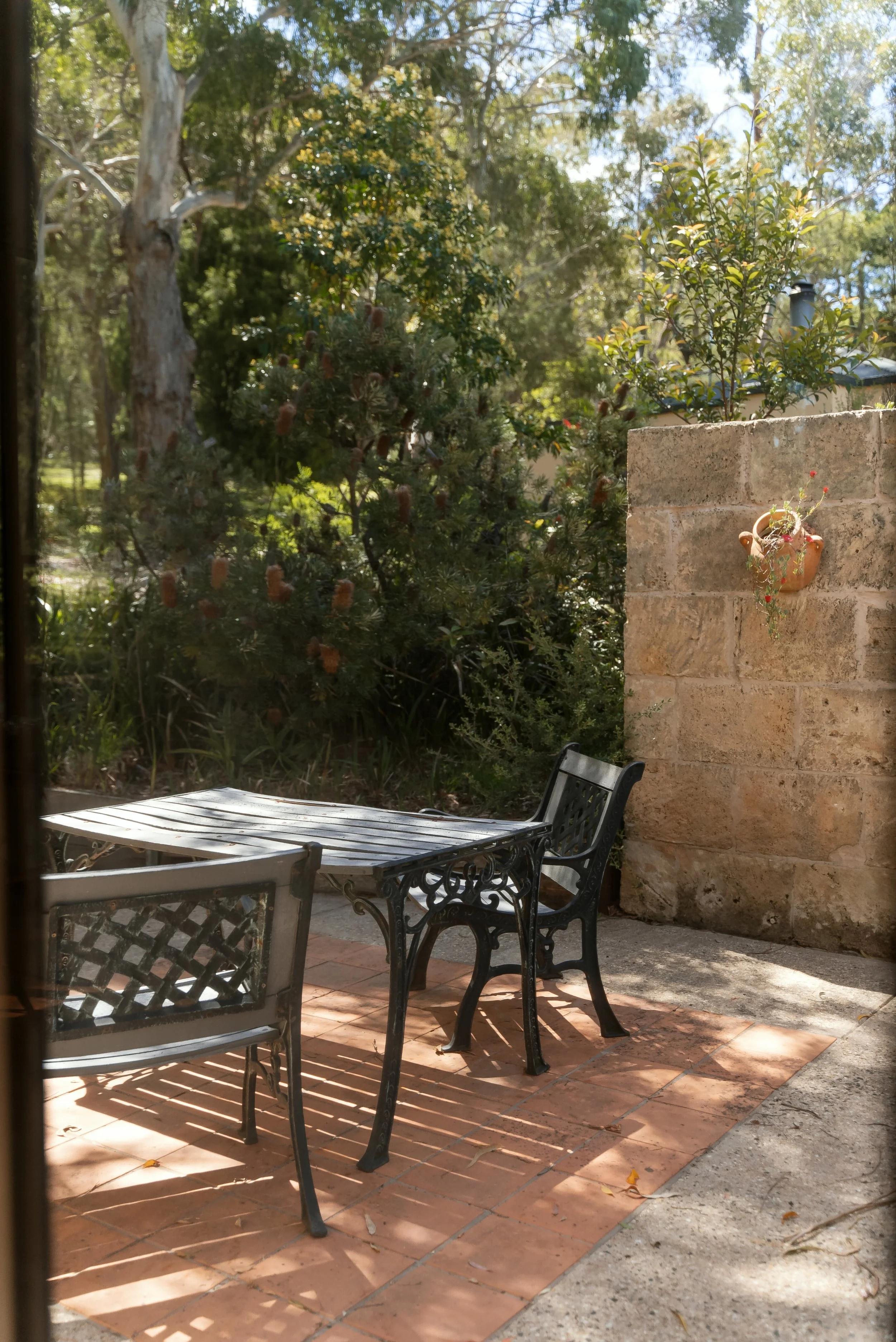A small outdoor patio with a metal table and chairs, surrounded by lush greenery and trees, with part of a stone wall with a hanging flower pot.