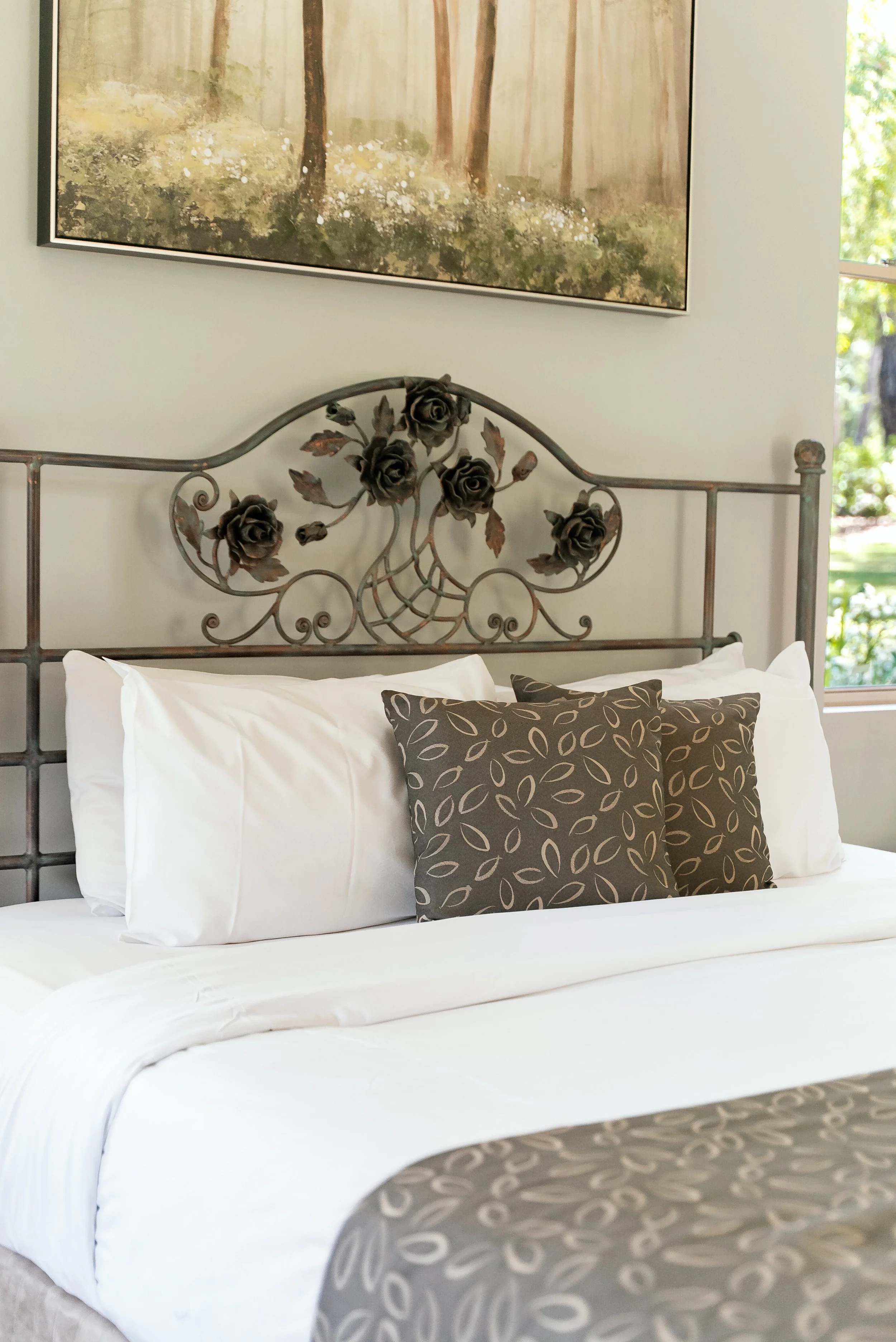 A bed with white bedding and decorative pillows featuring a brown and beige leaf pattern, set against a metal headboard with floral designs in a bedroom with a large wall art and window showing outdoor greenery.