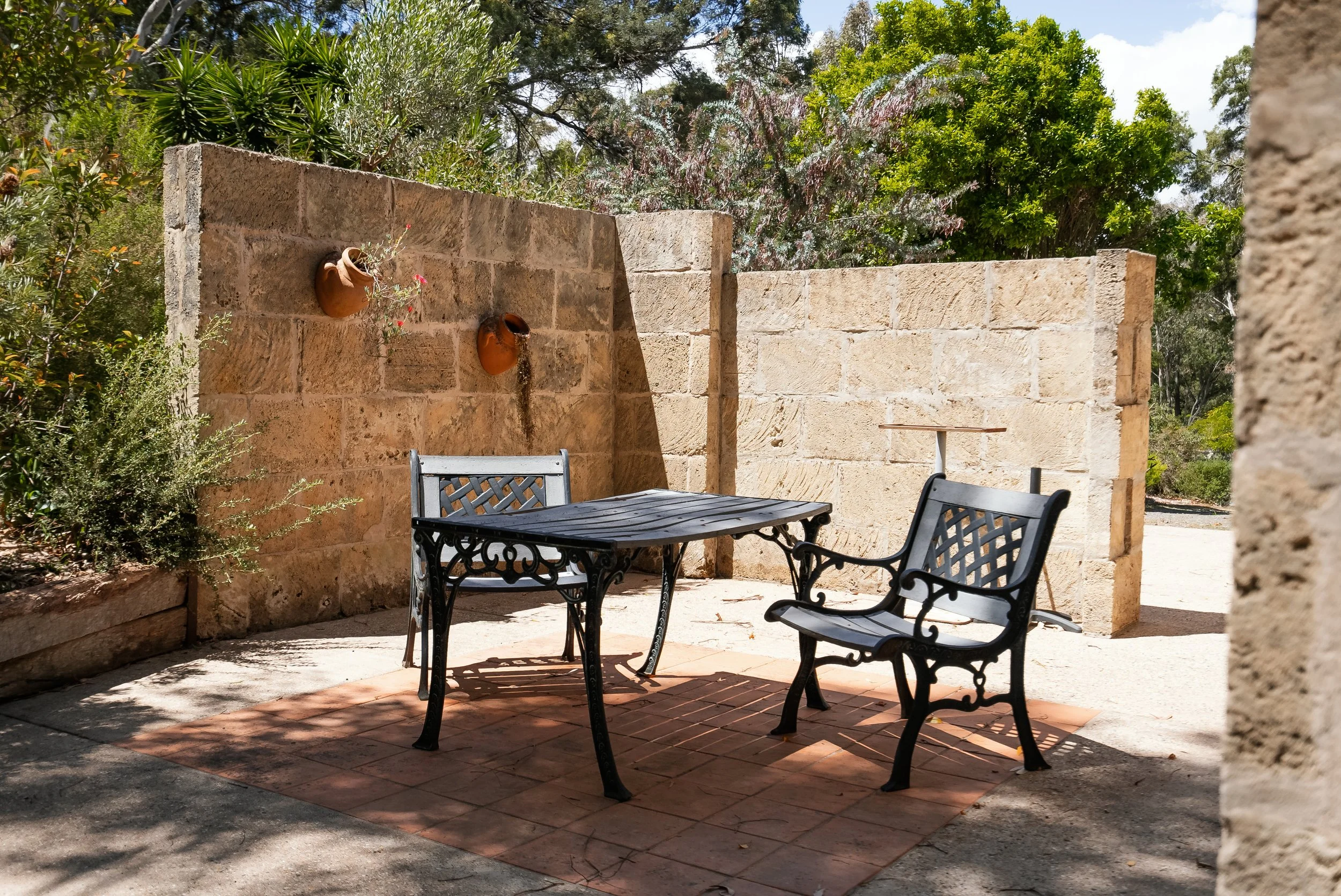 Outdoor patio with two metal benches and a table on a brick tile, surrounded by stone walls and greenery, with two hanging pottery planters on the back wall and trees in the background.