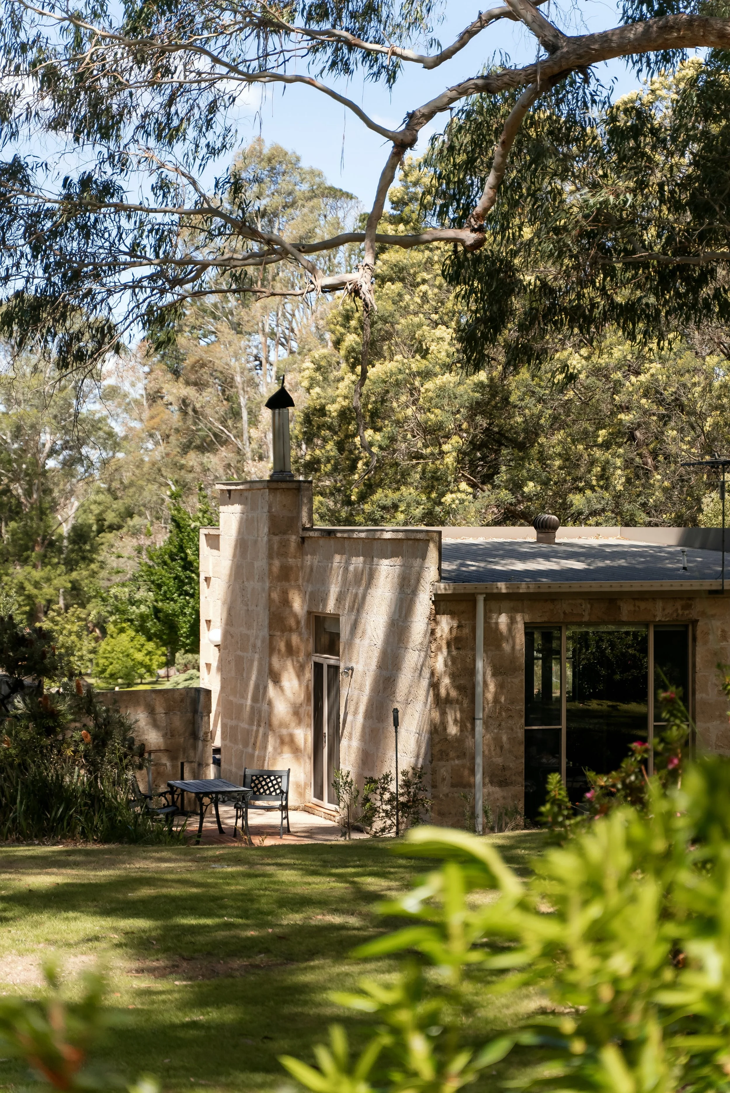 A stone house with a patio, surrounded by green grass and trees, under a partly cloudy sky.