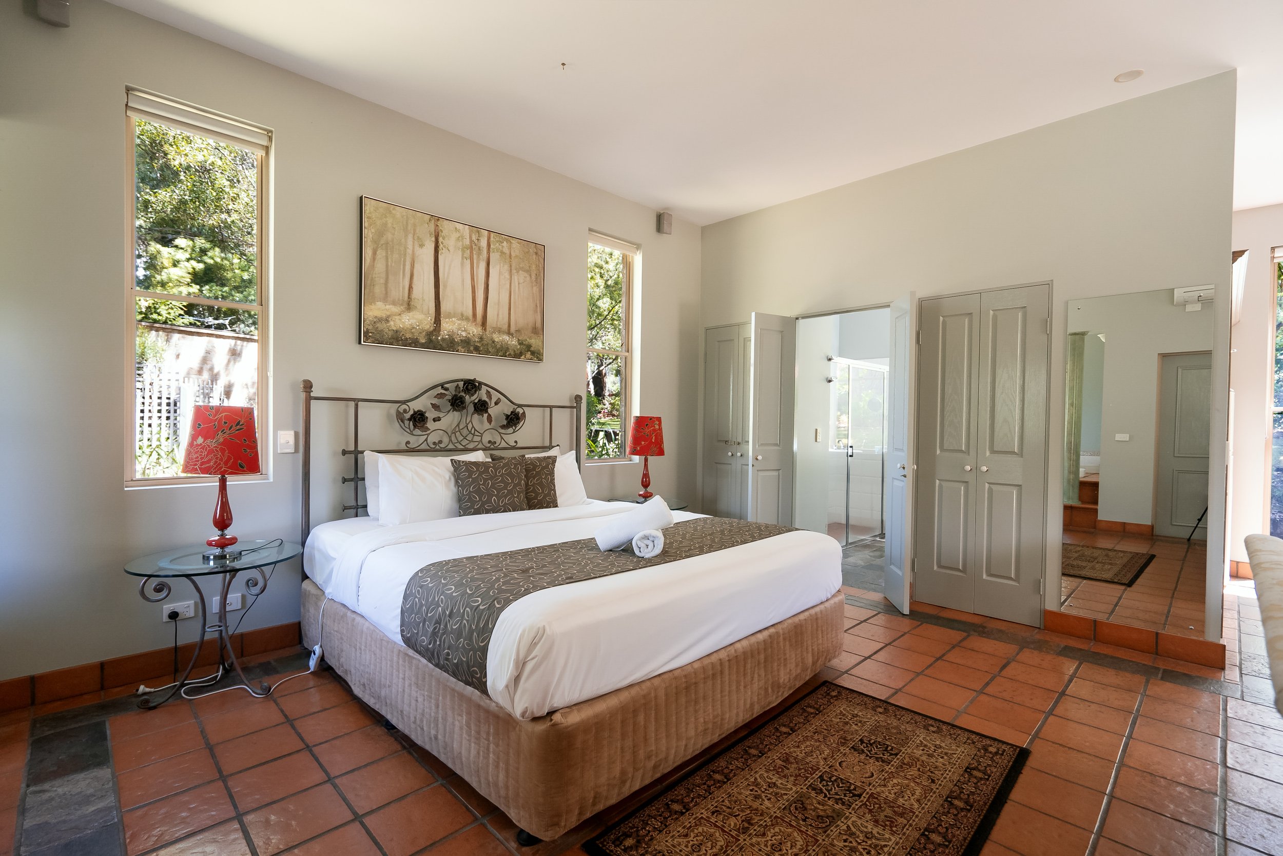 Bedroom with a bed, two red lamps, a painting of a forest, and windows showing garden view