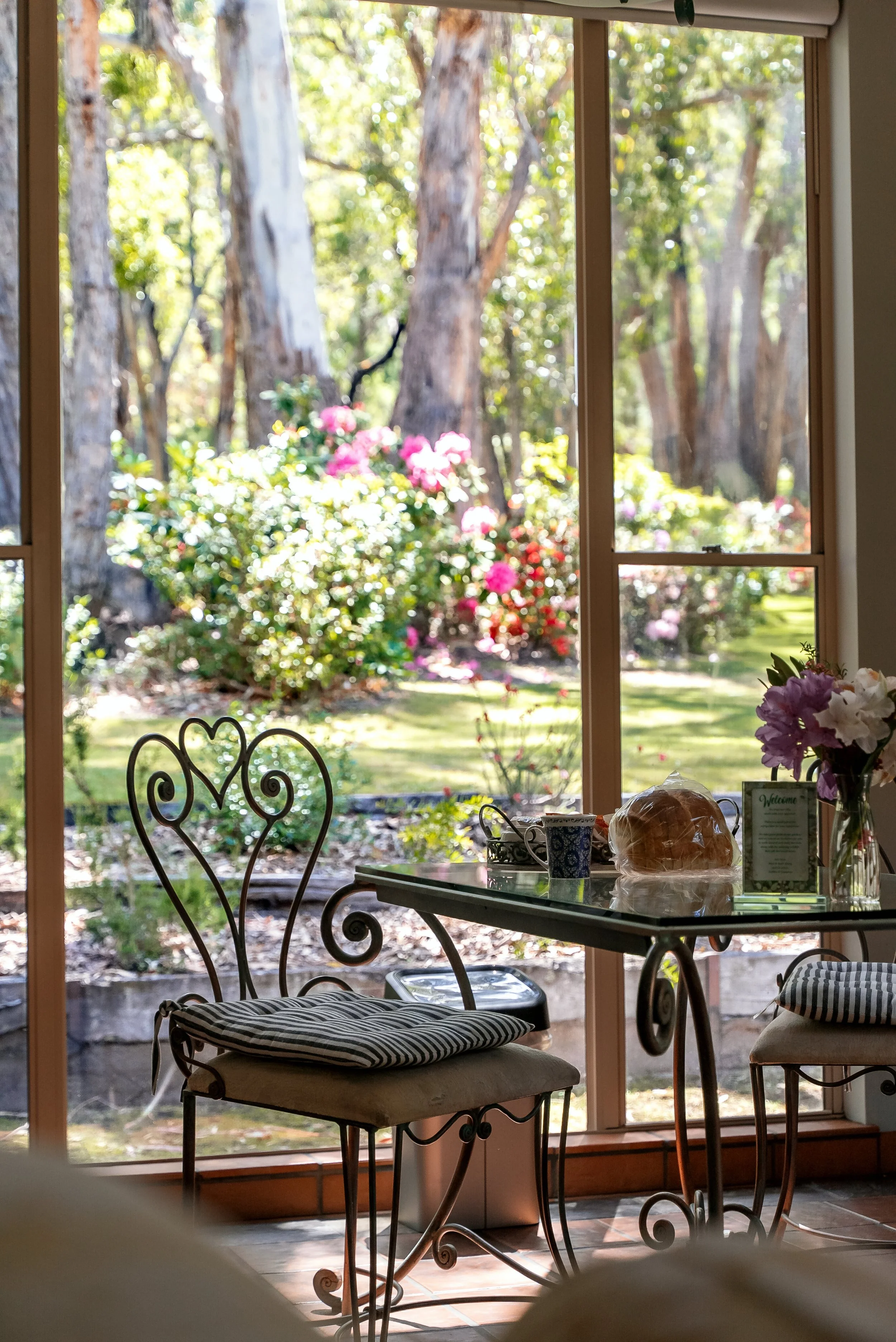Inside a cozy room with a glass table, wrought iron chairs with striped cushions, and a large window showing a garden with blooming flowers and trees outside.