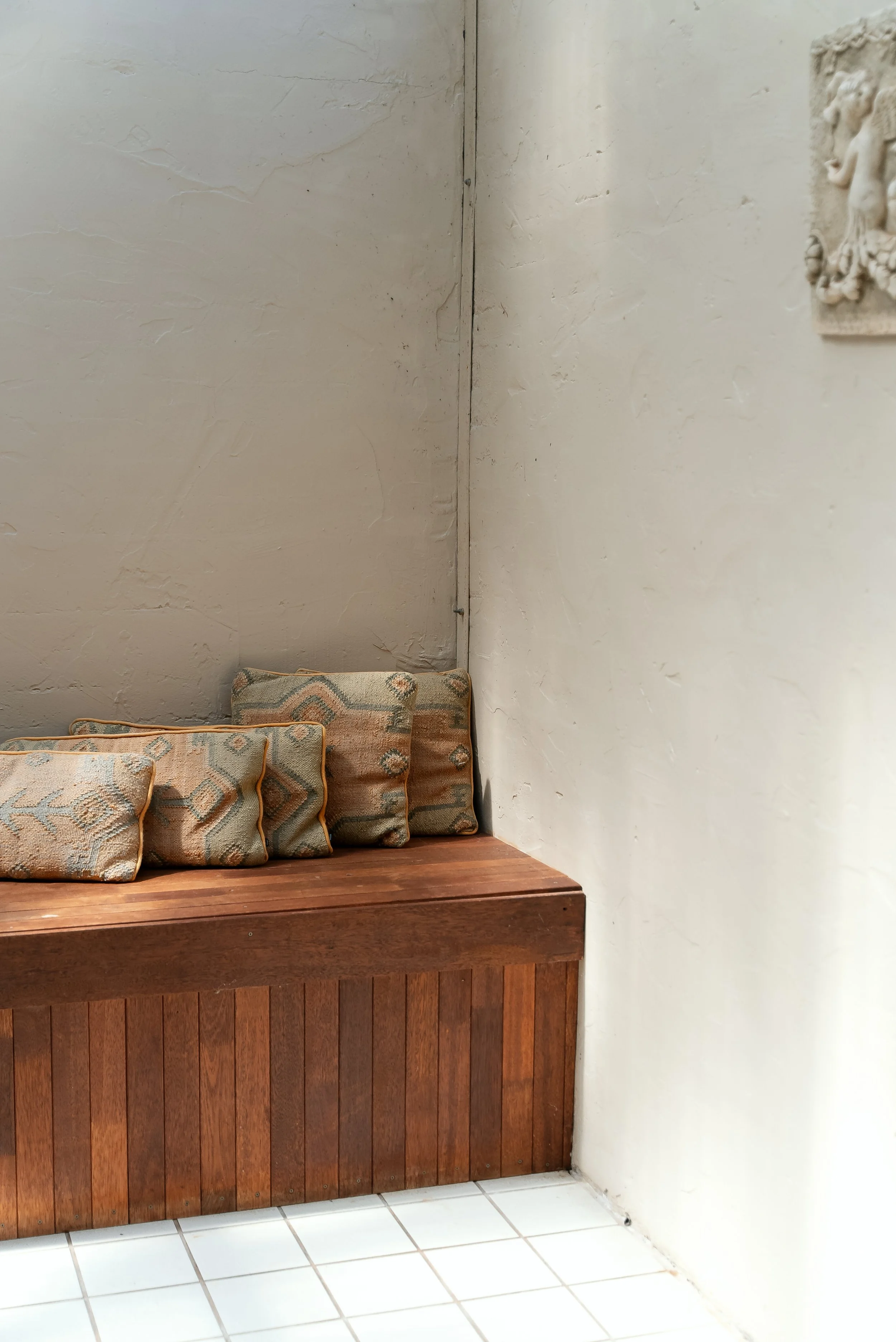 A wooden bench with five patterned cushions along the wall in an interior space with white tiled floor and textured light-colored walls.