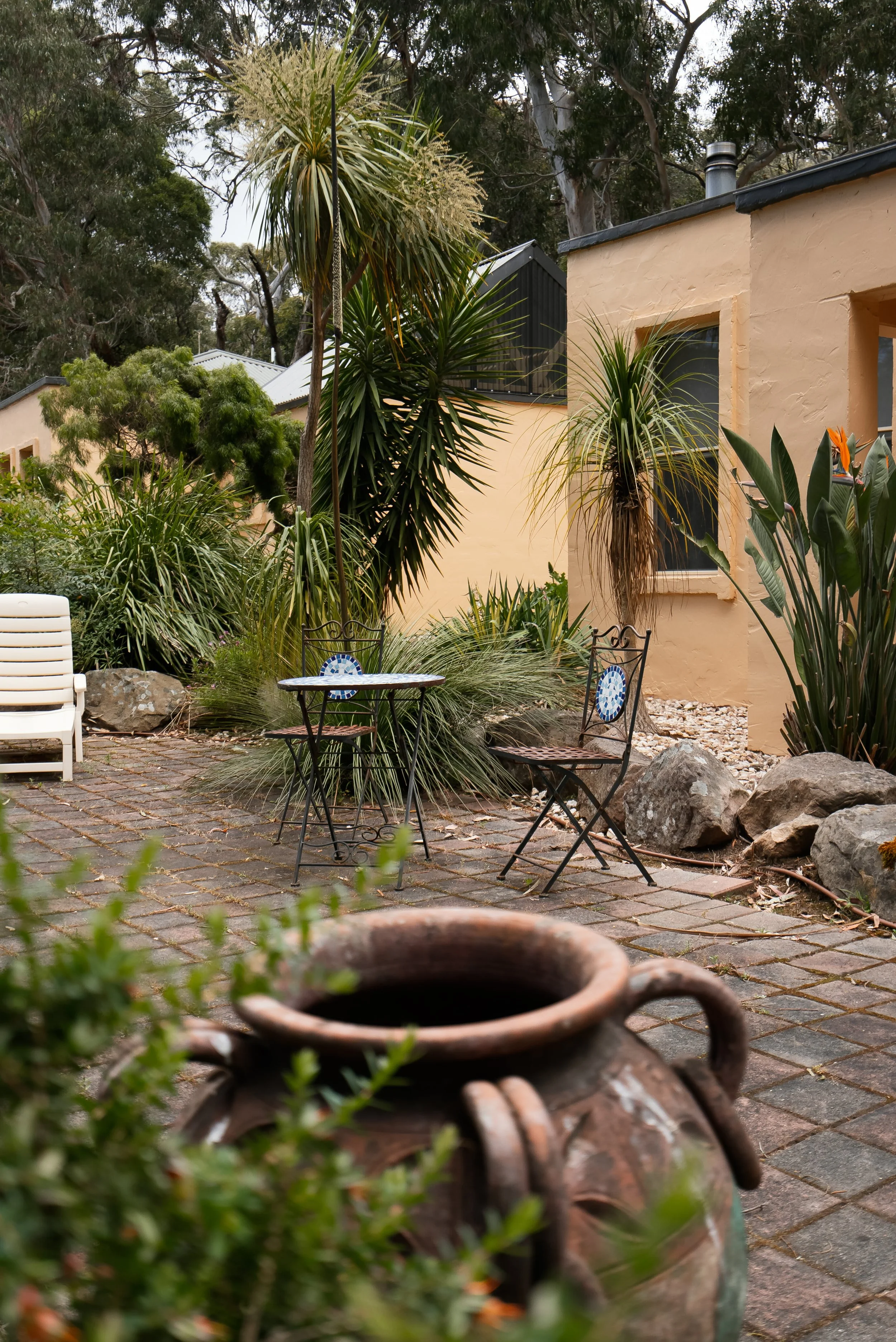 A cozy outdoor patio area with a small round table and two metal chairs with mosaic backs, surrounded by various lush tropical plants and rocks, with a yellow stucco house in the background.