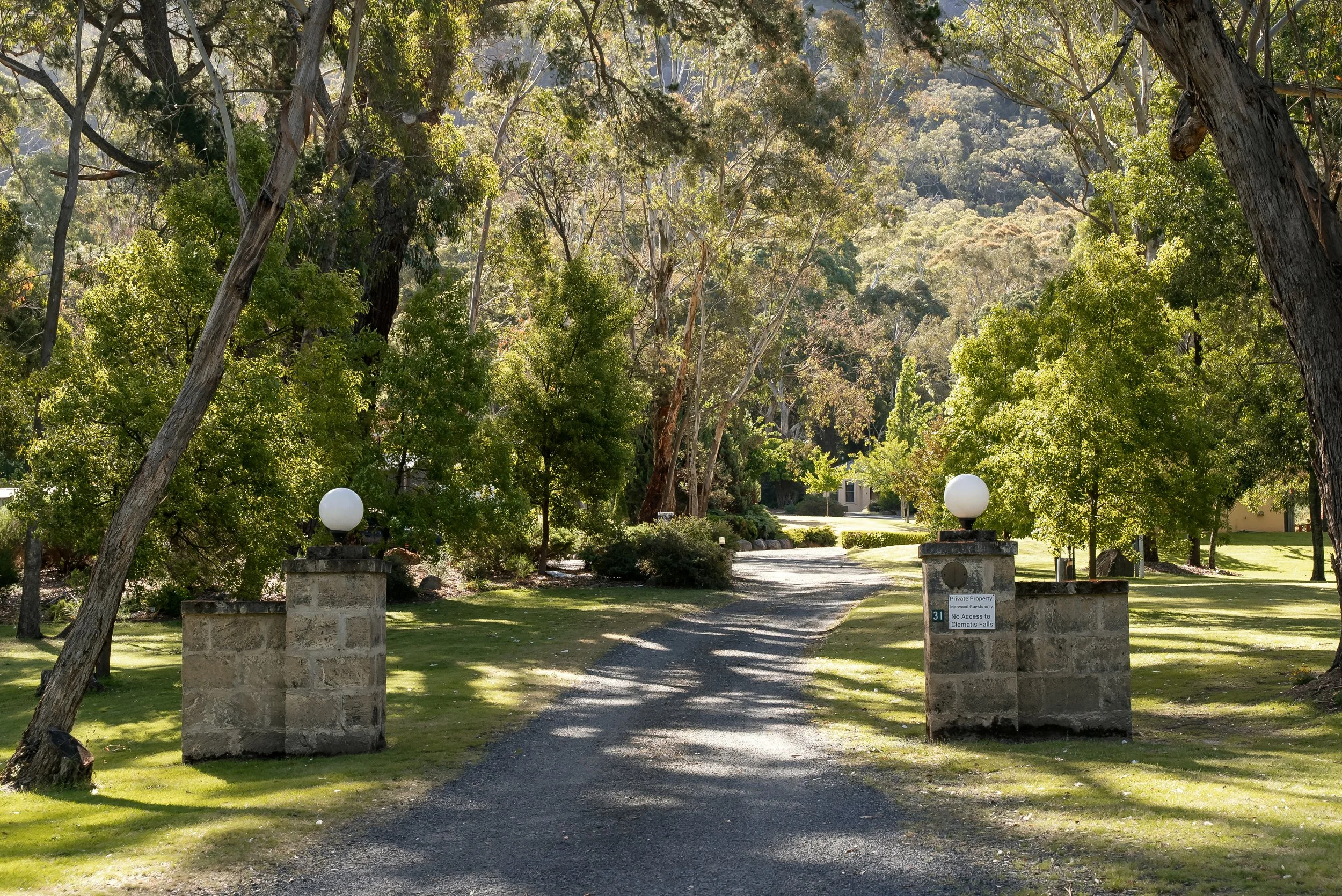 A gravel driveway leading into a lush, green park or estate with tall trees, stone pillars topped with white globes, and a small sign indicating no access to a creek or falls.