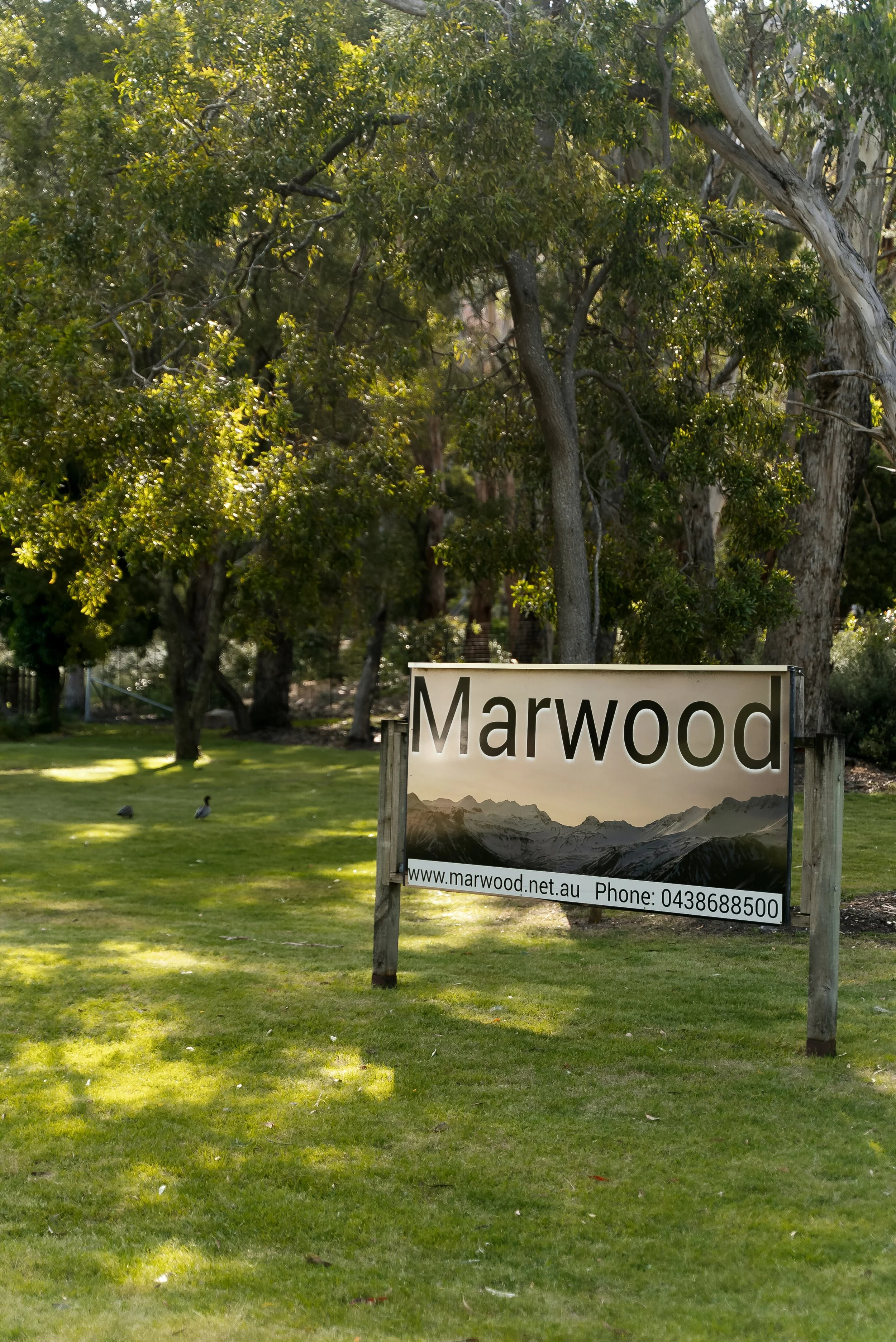 A sign that reads 'Marwood' with a mountain landscape background, located in a park with green grass and trees.