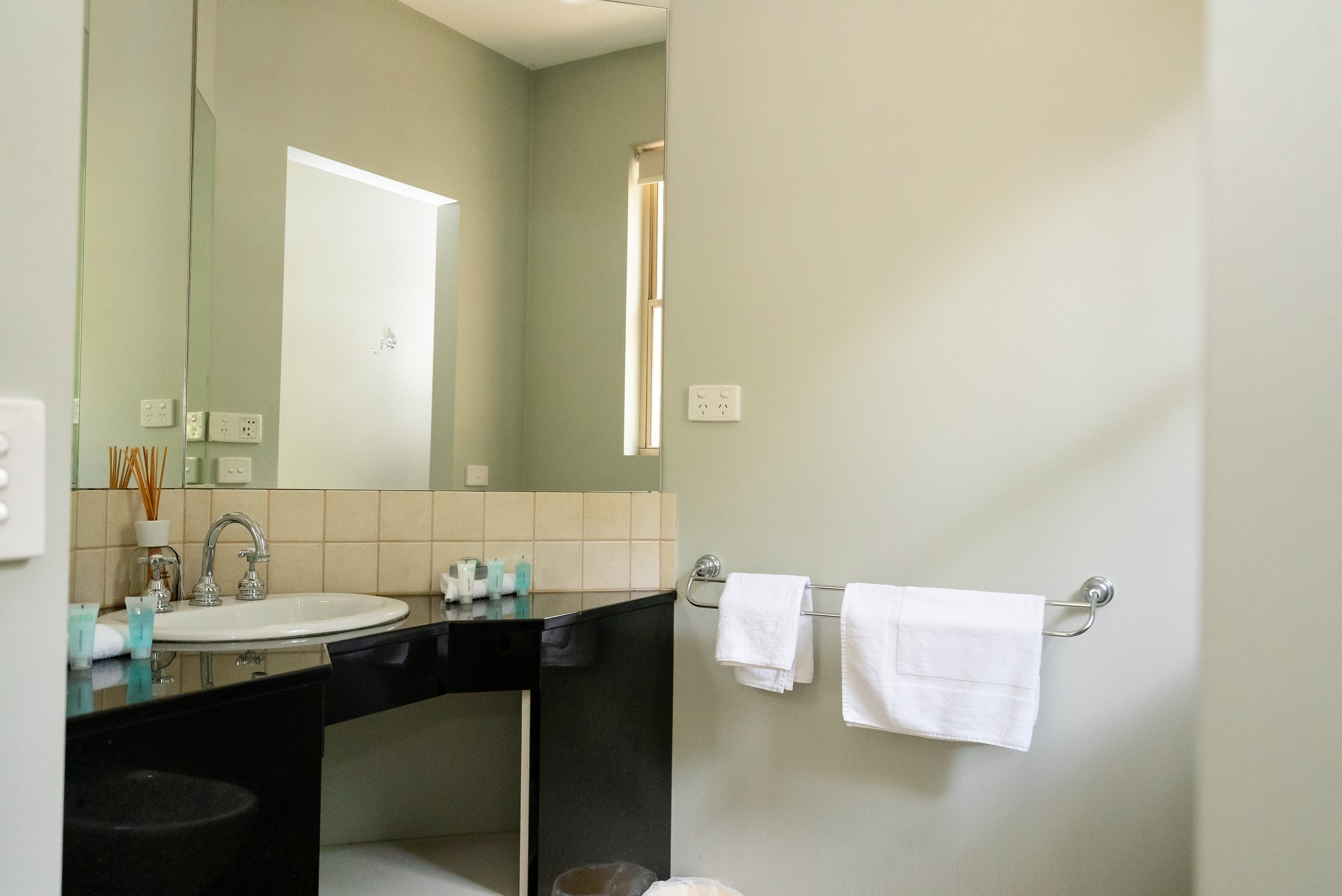 A bathroom with a mirror, sink, and a beige tile countertop. There are toiletries on the counter, a towel rack with two white towels, and an electrical outlet above the towel rack. A small narrow window is present, allowing natural light into the room.