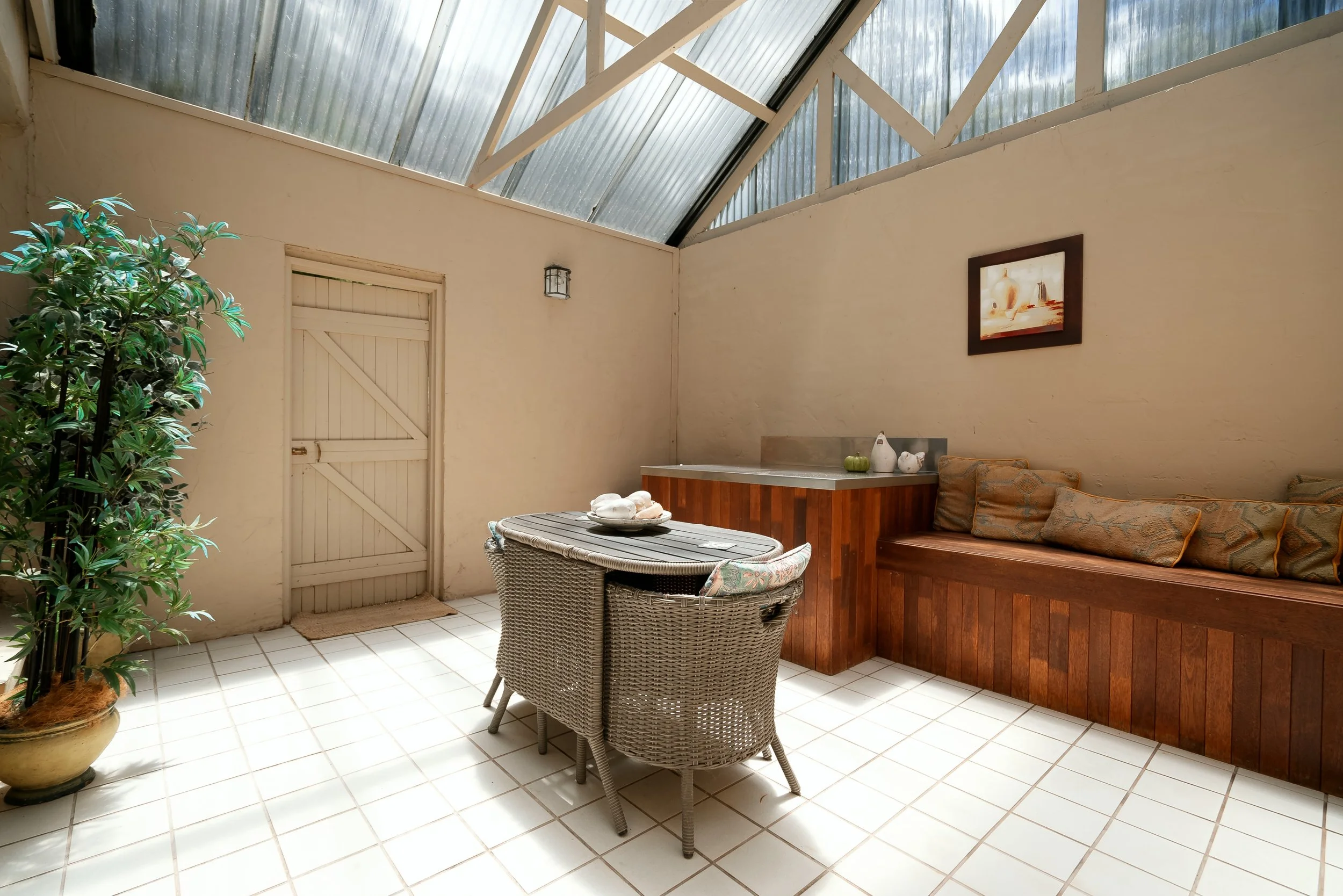 An indoor patio area with a glass ceiling, a white door on the left, a large potted plant, a round table with woven chairs, a built-in wooden bench with patterned cushions, a small countertop with decorative items, and a framed painting on the wall.