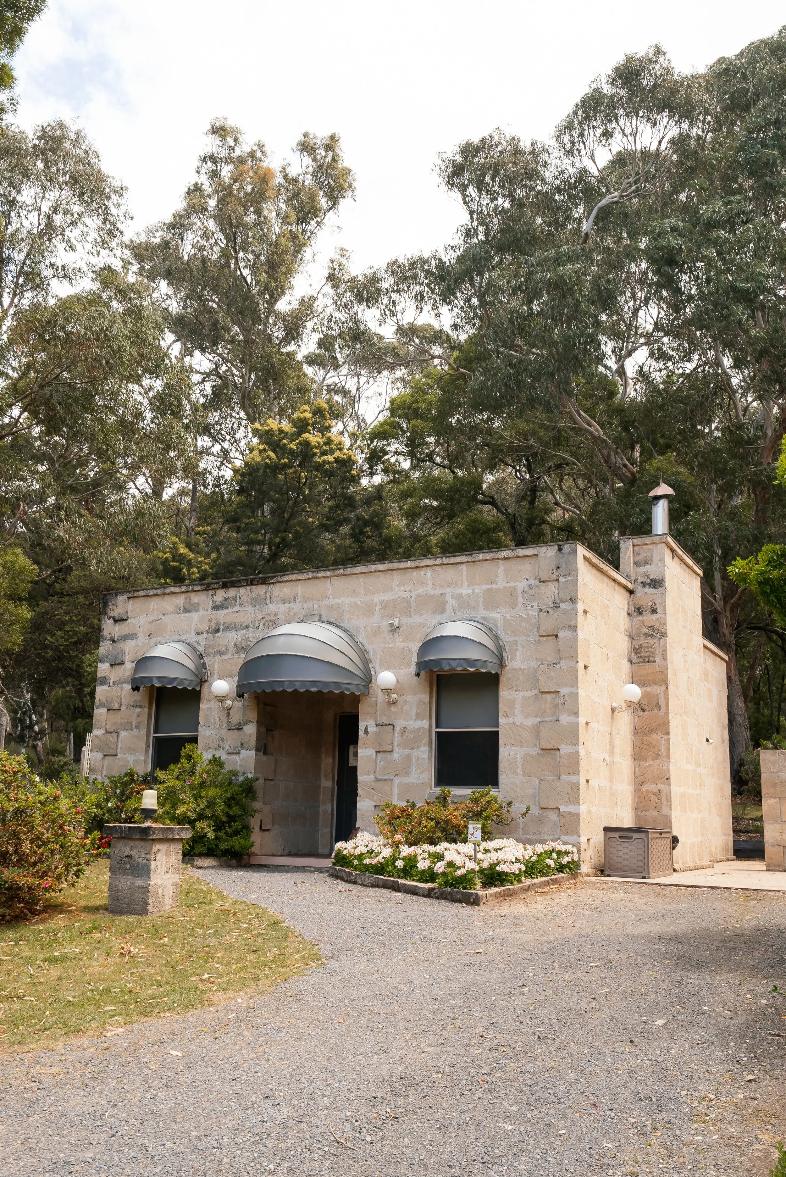 A small stone building with gray awnings over the windows and door, surrounded by greenery and flowers, with a gravel driveway in front.