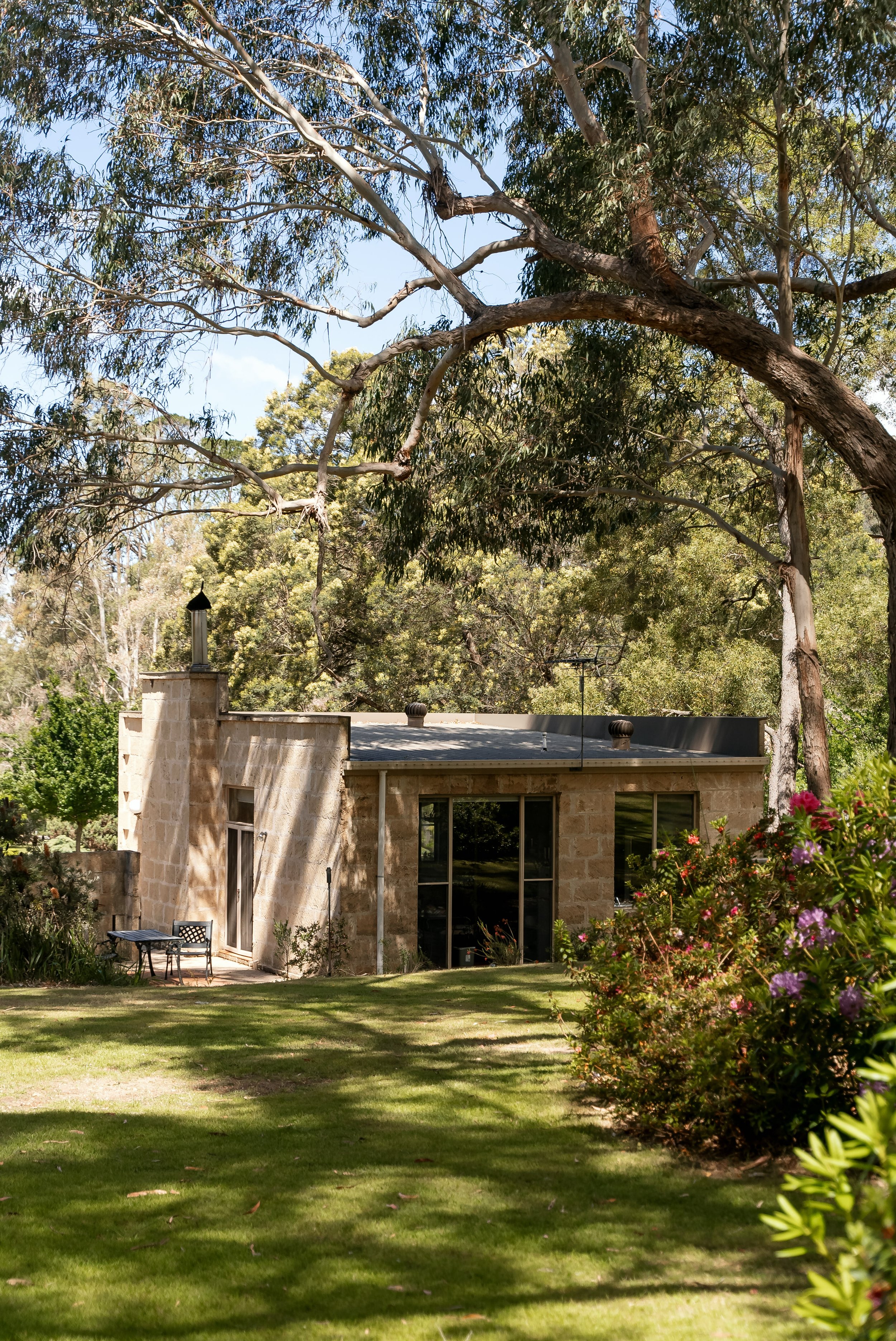 A stone house with large glass windows in a lush green yard, surrounded by trees and flowering bushes, under a bright blue sky.