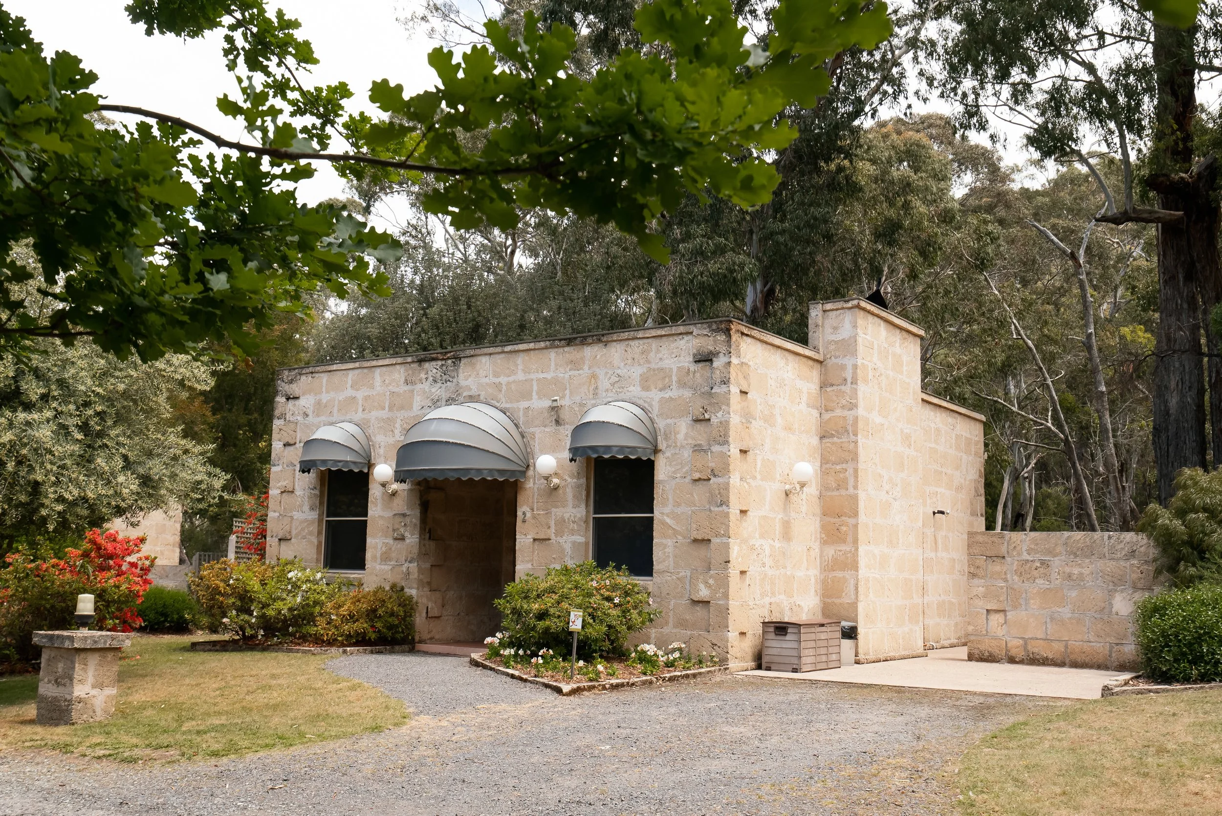 A stone building with three arched windows, covered entrances, and exterior wall-mounted lights, surrounded by greenery and trees.
