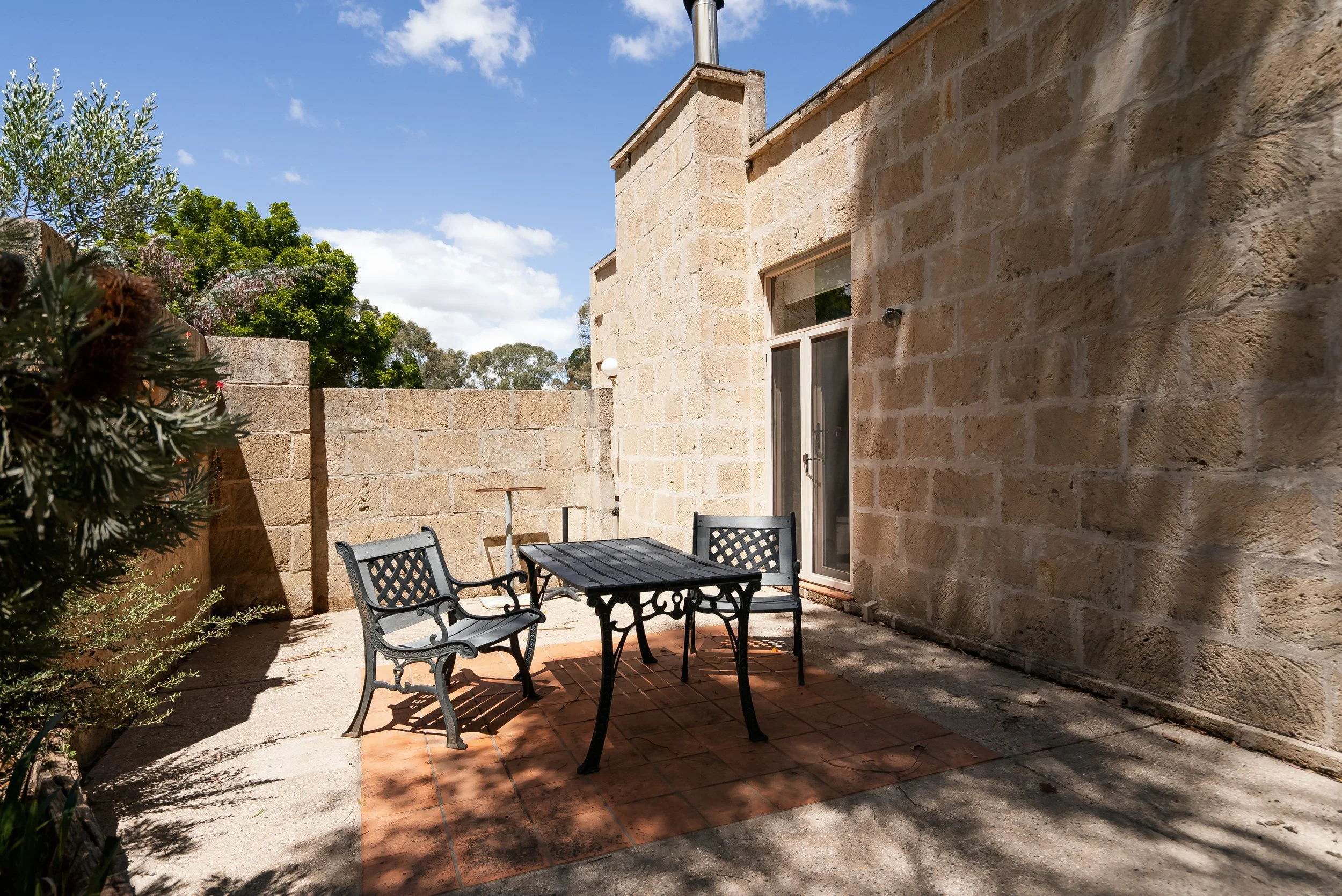 Outdoor patio with metal table and chairs, brick and stone wall, sliding glass door, and trees in the background under partly cloudy sky.