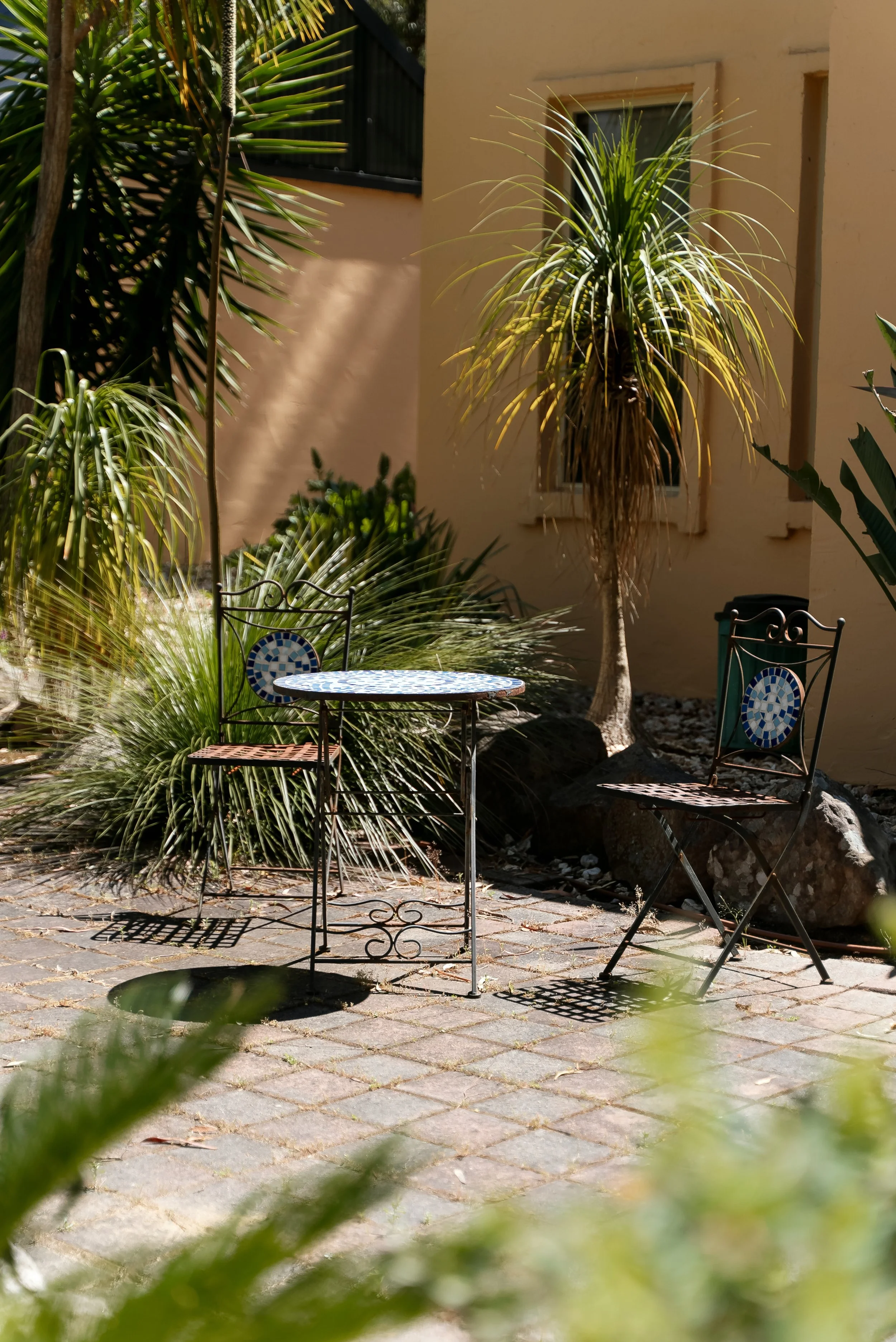A small outdoor patio with a round mosaic table and two wrought iron chairs, surrounded by tropical plants and rocks, next to a beige stucco wall with a window.