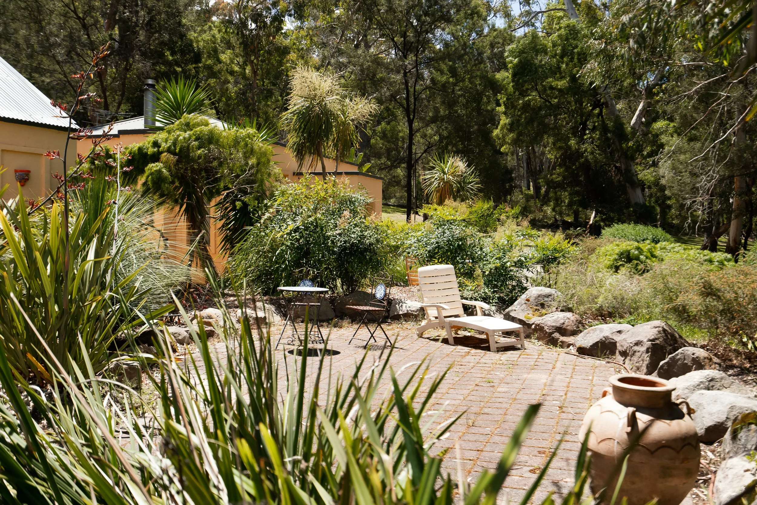 A backyard patio with a white lounge chair, a small black metal table with two matching chairs, surrounded by various green plants, trees, and rocks, with a tan house in the background and a paved brick floor.