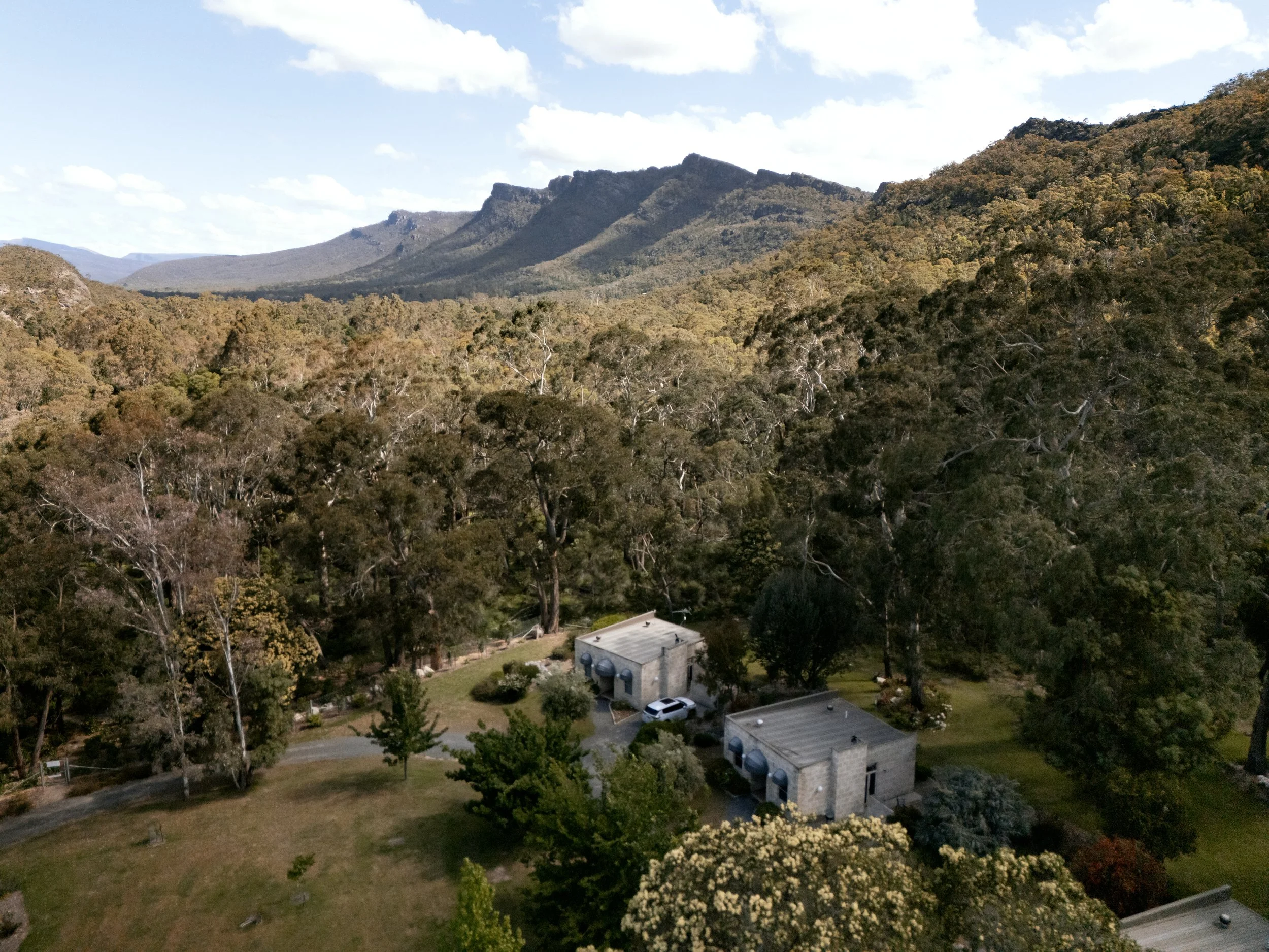 Aerial view of a rural area with two modern houses surrounded by trees and greenery, with mountains in the background, under a partly cloudy sky.