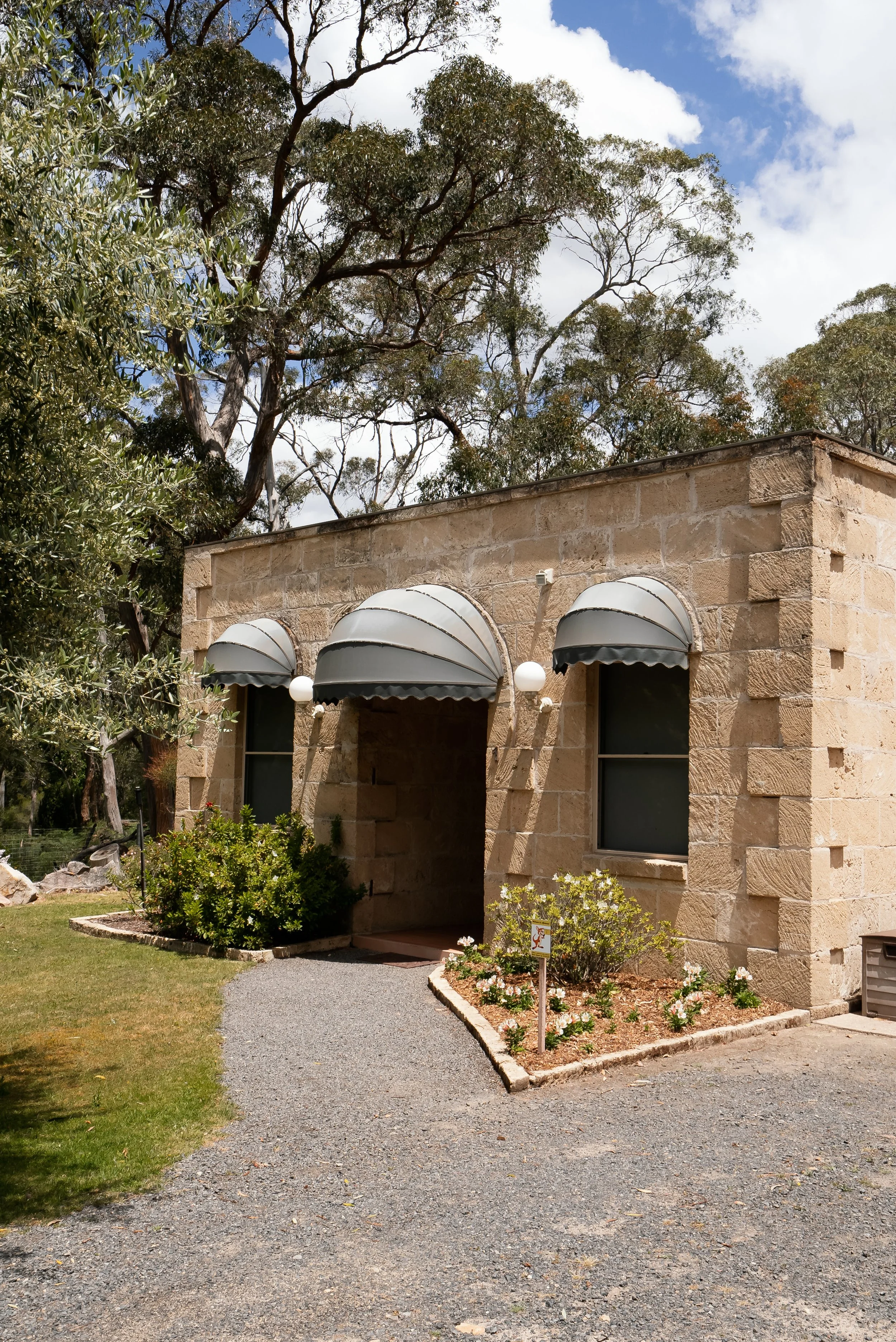 A stone building with two windows and small rounded awnings, surrounded by trees and flowerbeds, under a partly cloudy sky.