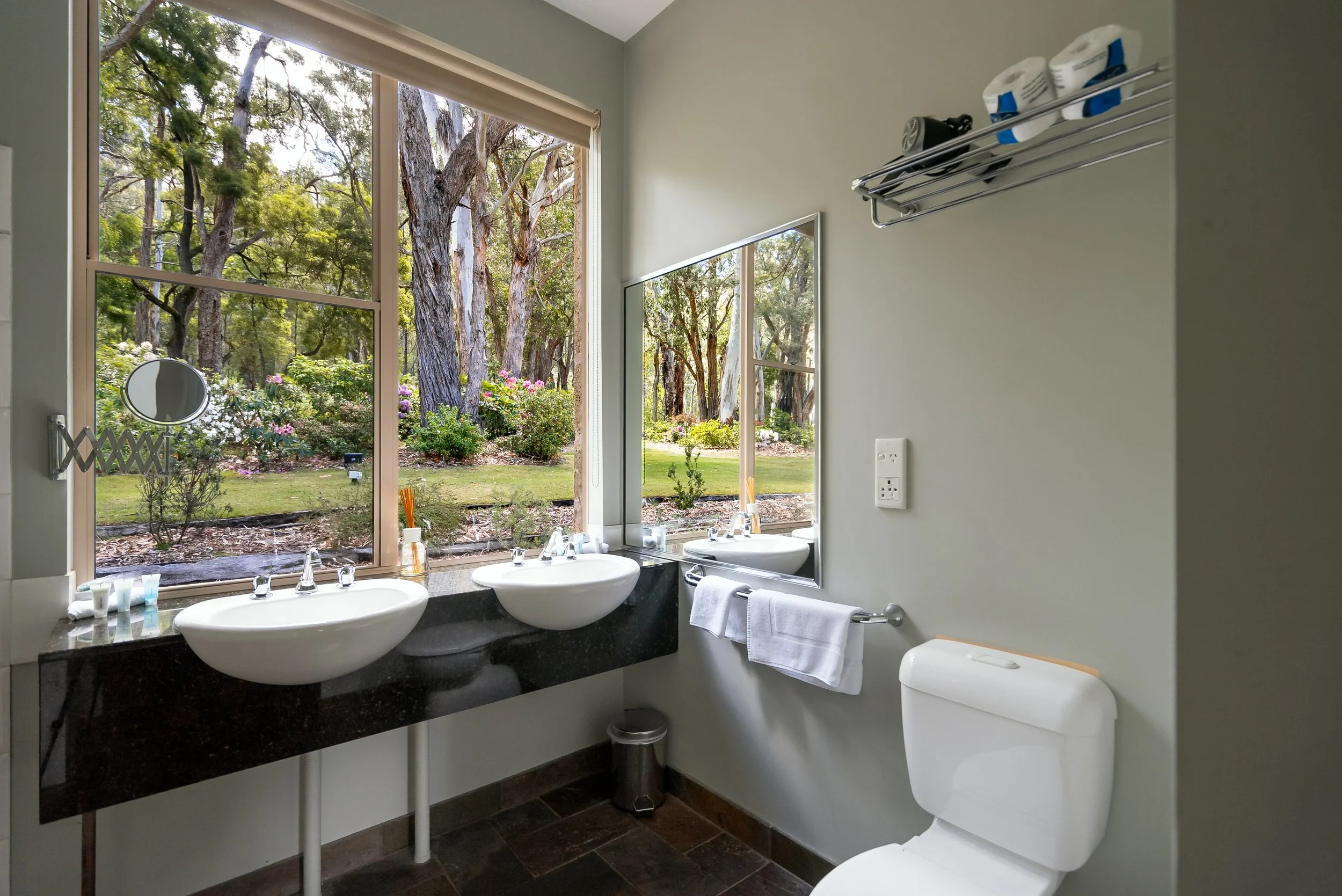 Bathroom with two sinks, a toilet, and large windows showing a wooded outdoor scene with trees and bushes.