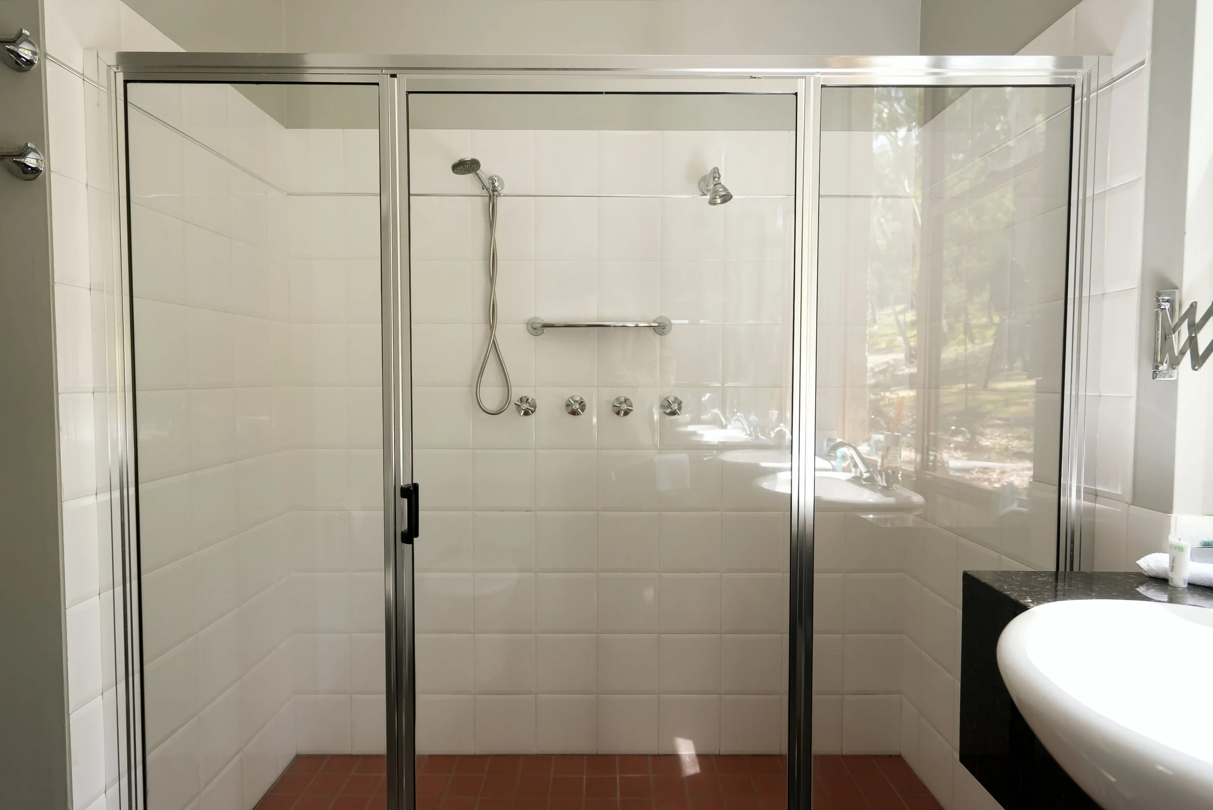 Empty glass shower enclosure with metal frame, white tiled walls, a hand-held showerhead, a small built-in shelf, and four control knobs. Part of a white sink and black countertop are visible on the right, with a window showing outdoor trees and cars in the background.