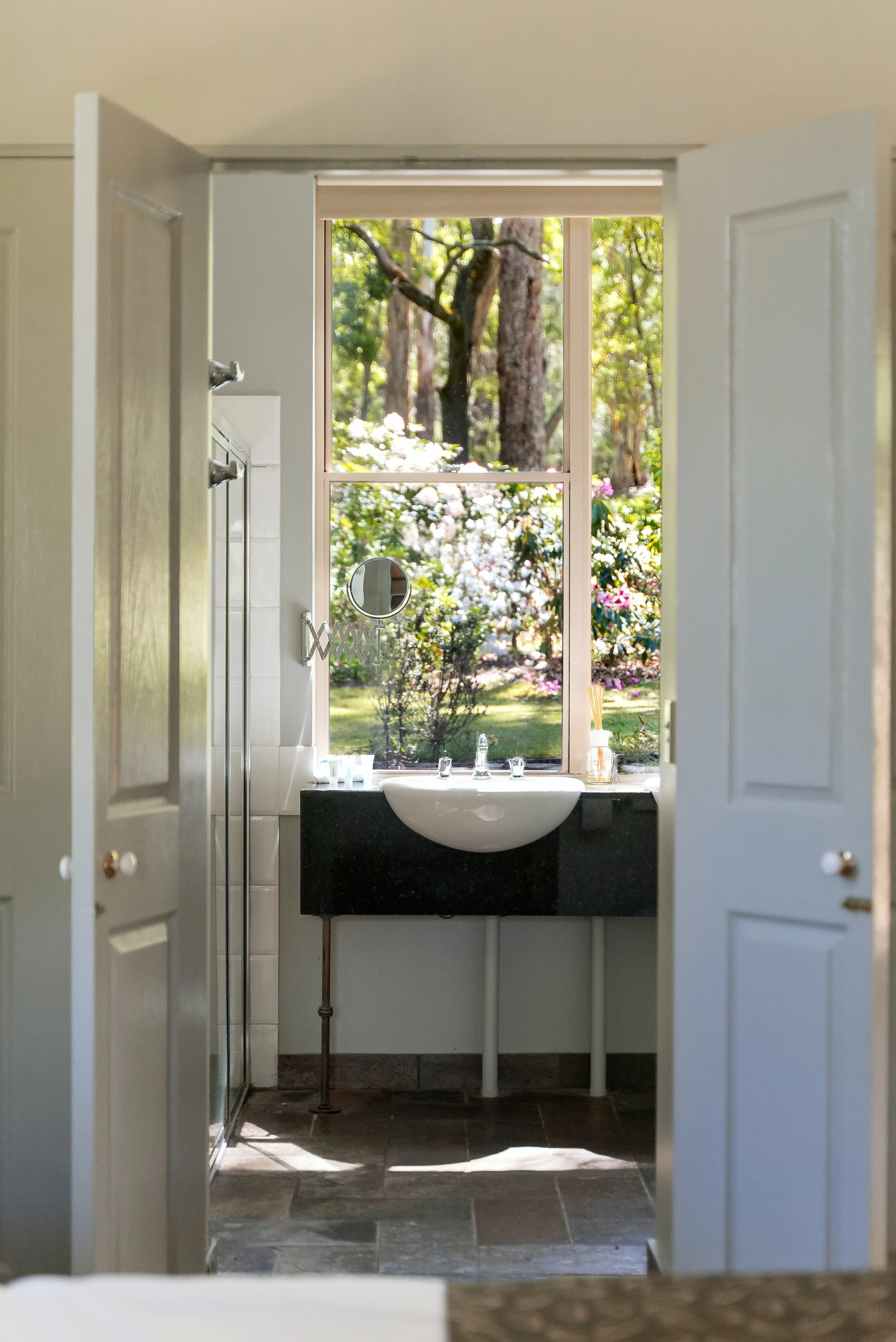 Bathroom with a large window showing a garden with trees and flowering bushes, a white vessel sink on a black countertop, a small round mirror, and a shower door.