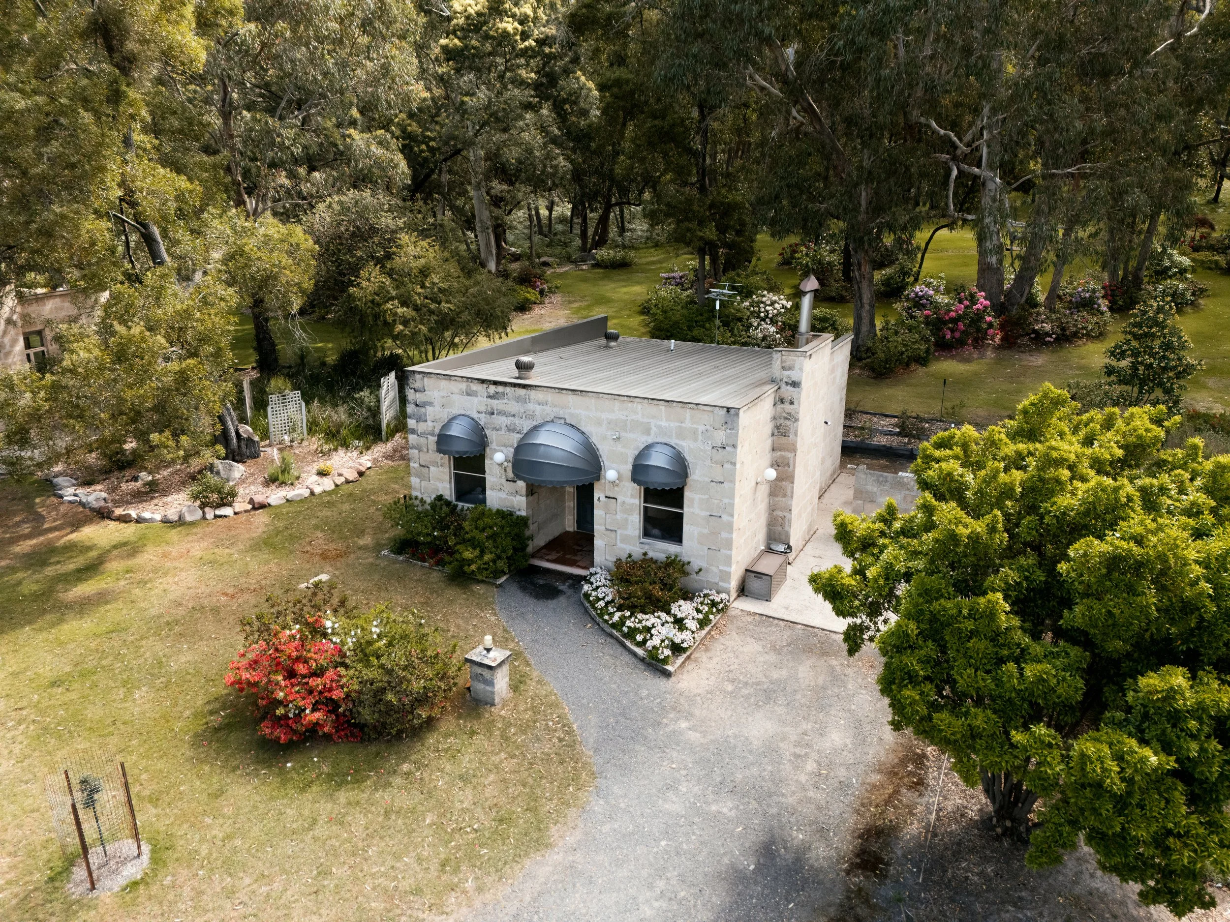 A small building with stone walls and three black awnings on the front, surrounded by a grassy yard with bushes and flowers, and tall trees in the background.