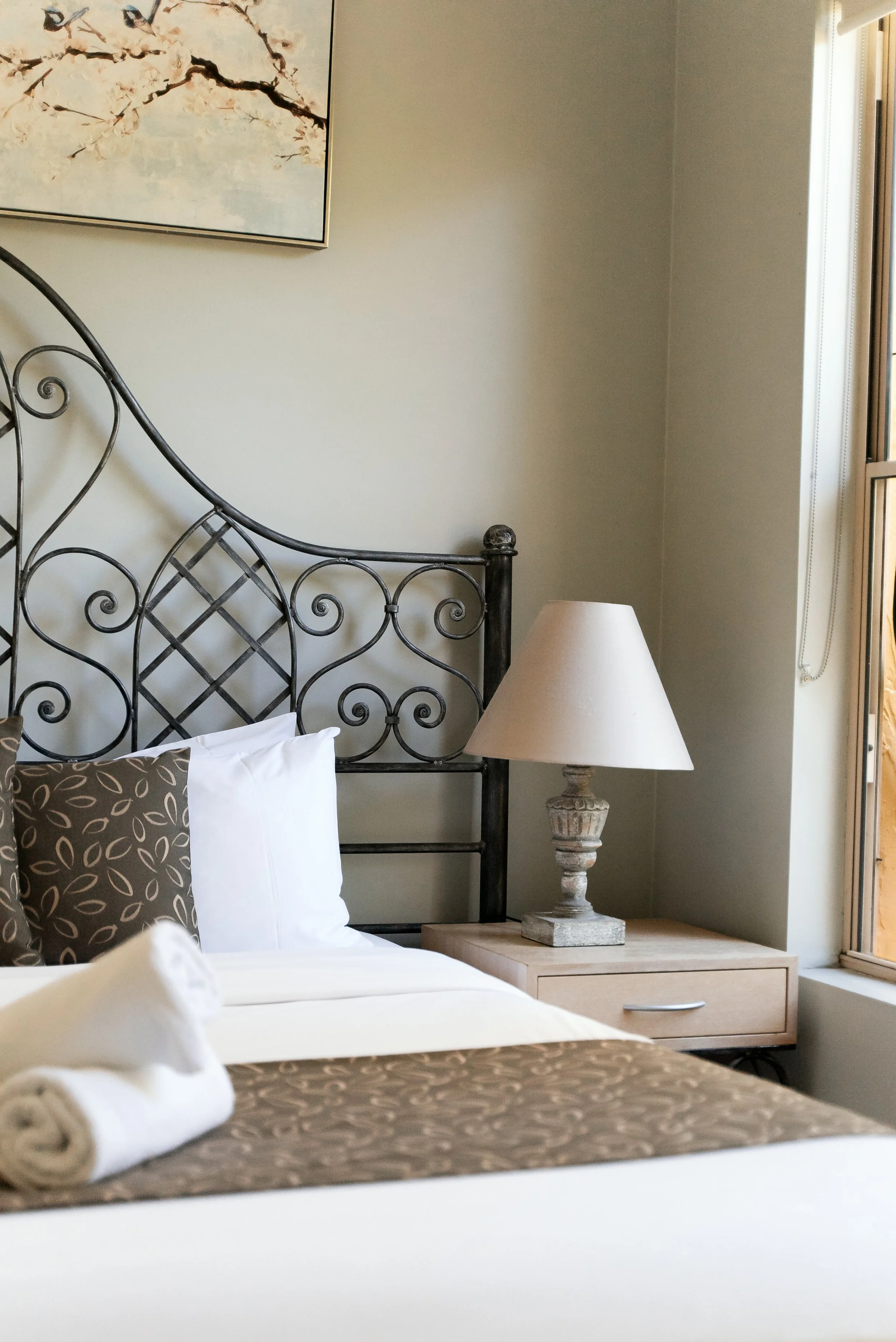 A neatly made bed with white sheets, brown patterned pillows, and a rolled white towel on top. A vintage-style lamp sits on a wooden nightstand beside the bed, with a window to the right and an abstract painting of a flowering branch hanging on the wall above.