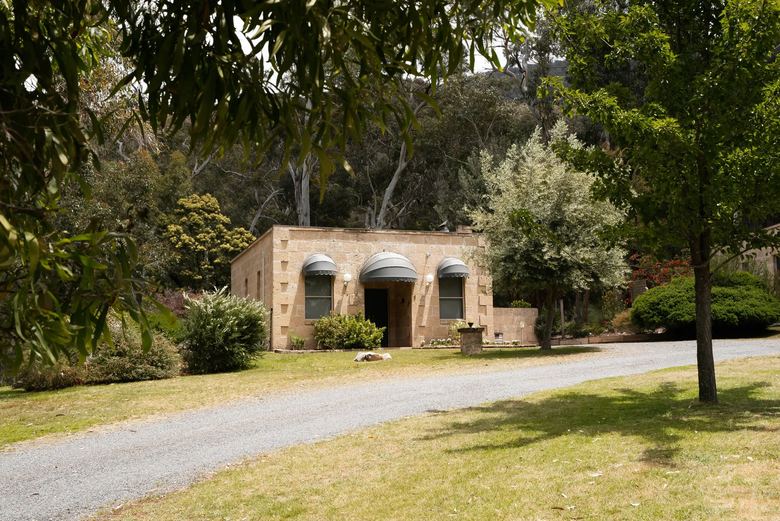 A small stone house with three arching awnings over the windows and door, surrounded by trees and greenery, with a gravel driveway in front.