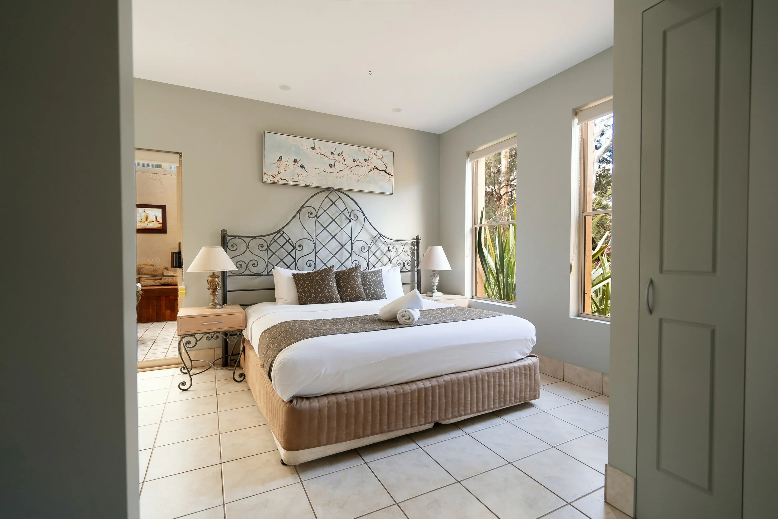 A bedroom with a bed, bedside tables, lamps, and windows showing greenery outside.